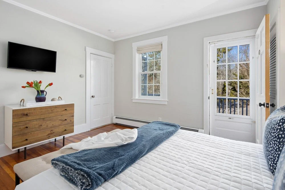 Bright bedroom with white walls, wooden floor, and large windows overlooking trees. A bed with a blue blanket and white sheets is in the foreground. A wall-mounted flat-screen TV and a wooden dresser with a vase of red tulips are on the left.