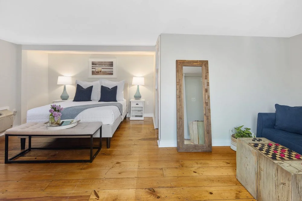 A bedroom with a white bed, navy pillows, two bedside lamps, a wooden floor, a mirror, a blue sofa, and a coffee table with a chess set.
