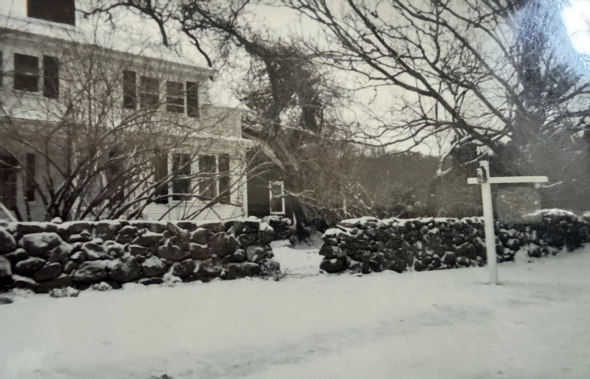 A snow-covered scene featuring a stone wall in the foreground, leafless trees with snow on their branches, and a stone house with multiple windows in the background. There is also a white signpost on the right side of the scene.