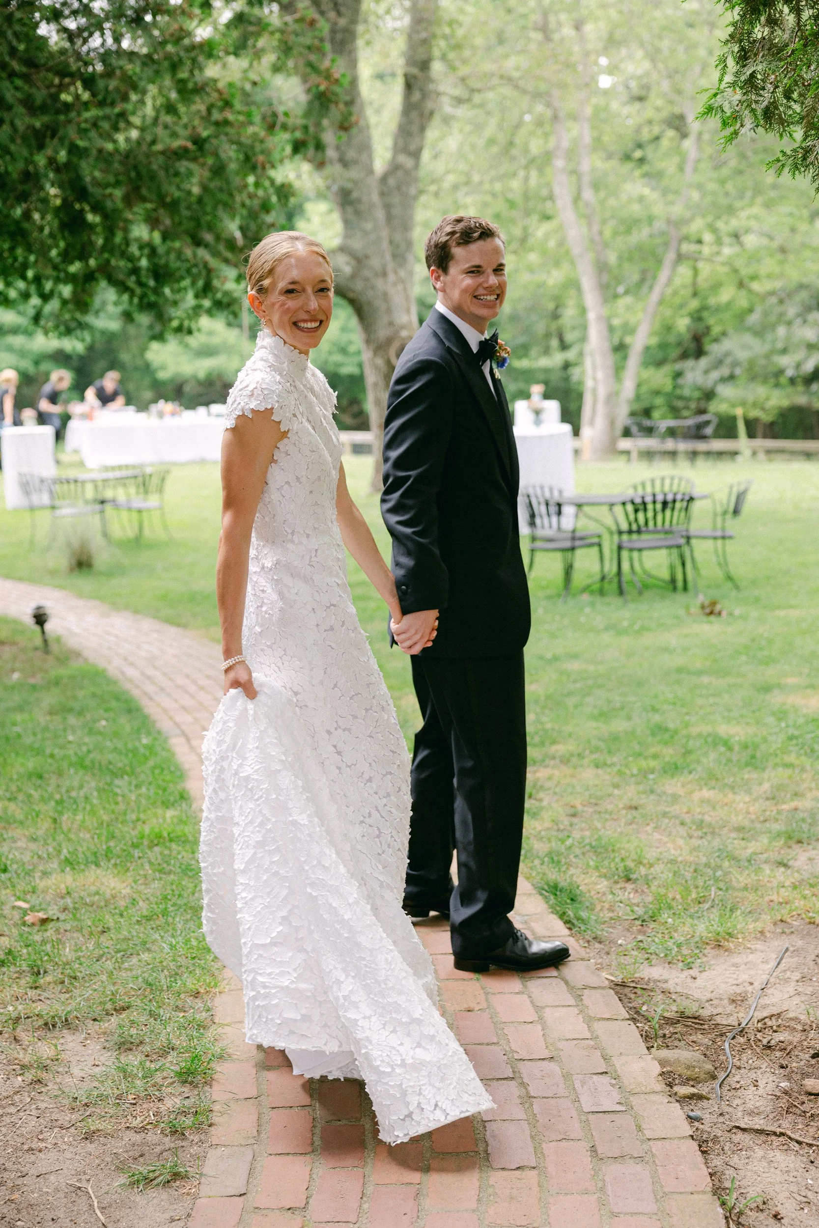 A smiling bride and groom holding hands on a brick pathway outdoors, with wedding reception tables and people in the background surrounded by trees during a wedding at Lambert's Cove Inn on Martha's Vineyard.