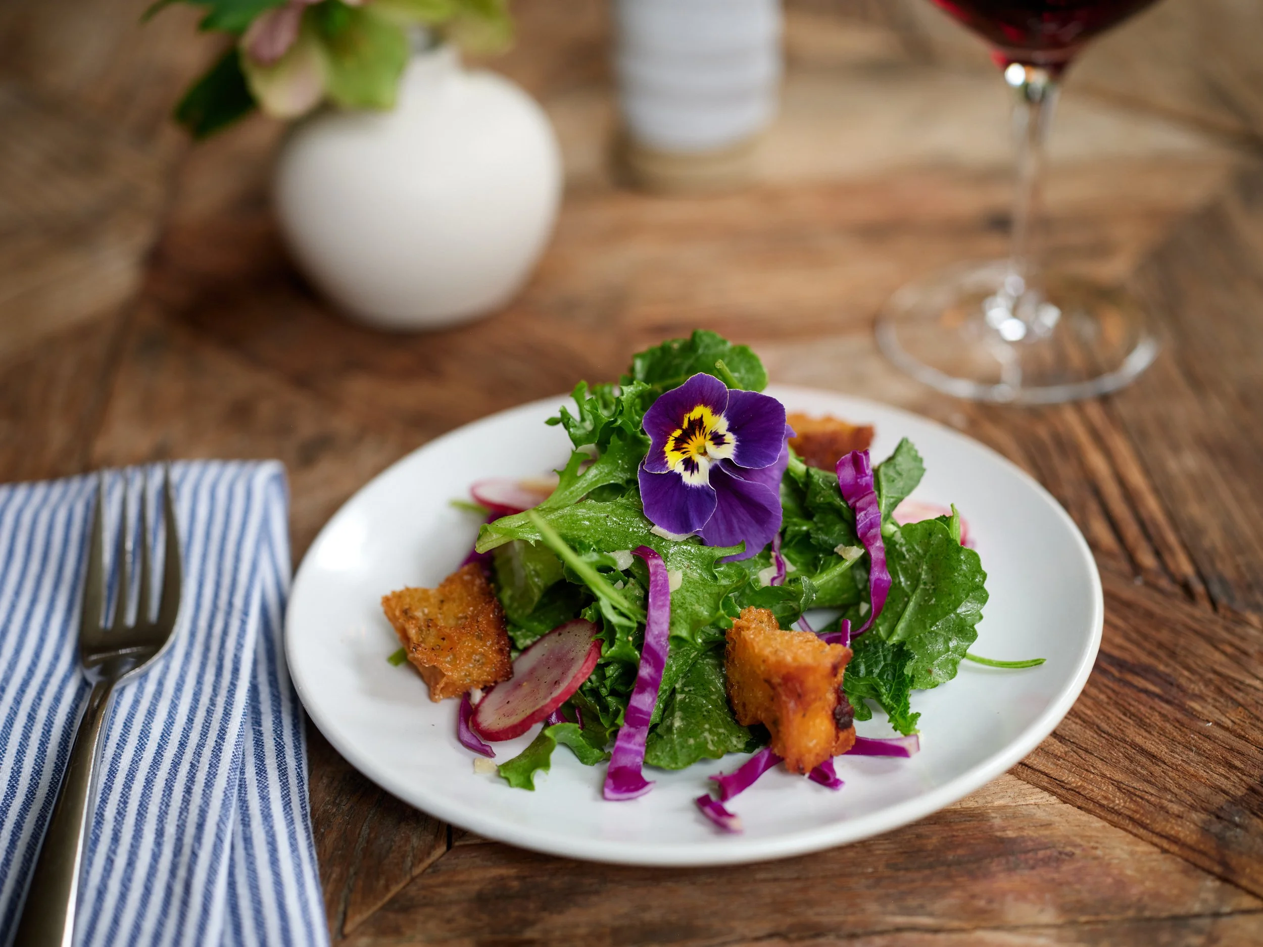A white plate with a salad containing greens, radish slices, and purple edible flowers, set on a wooden table with a blue-striped napkin and silver fork, with a glass of red wine and a small white vase in the background at Restaurant Foster.
