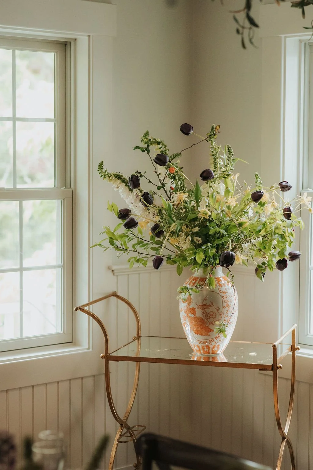 A floral arrangement with dark purple tulips, white lilies, and greenery in a decorative vase on a glass and gold table inside a room with large windows.