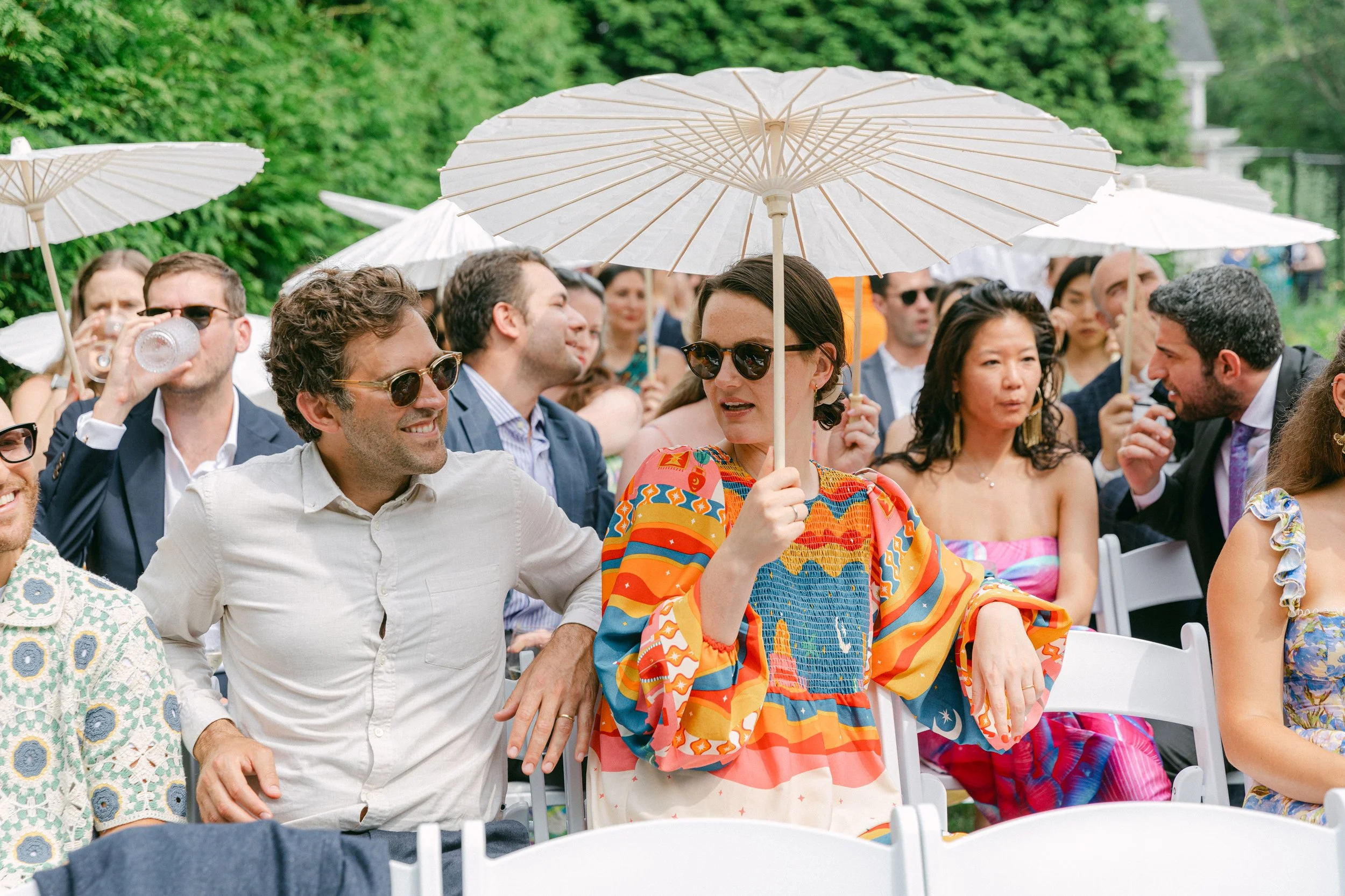 People gathered outdoors during a wedding at Lambert's Cove Inn on Martha's Vineyard, sitting on white chairs, some holding umbrellas, with green trees in background.