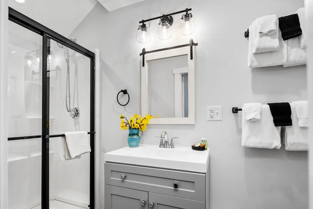 Modern bathroom with a glass shower, white vanity, framed mirror, black and white towels, and a vase with yellow flowers.