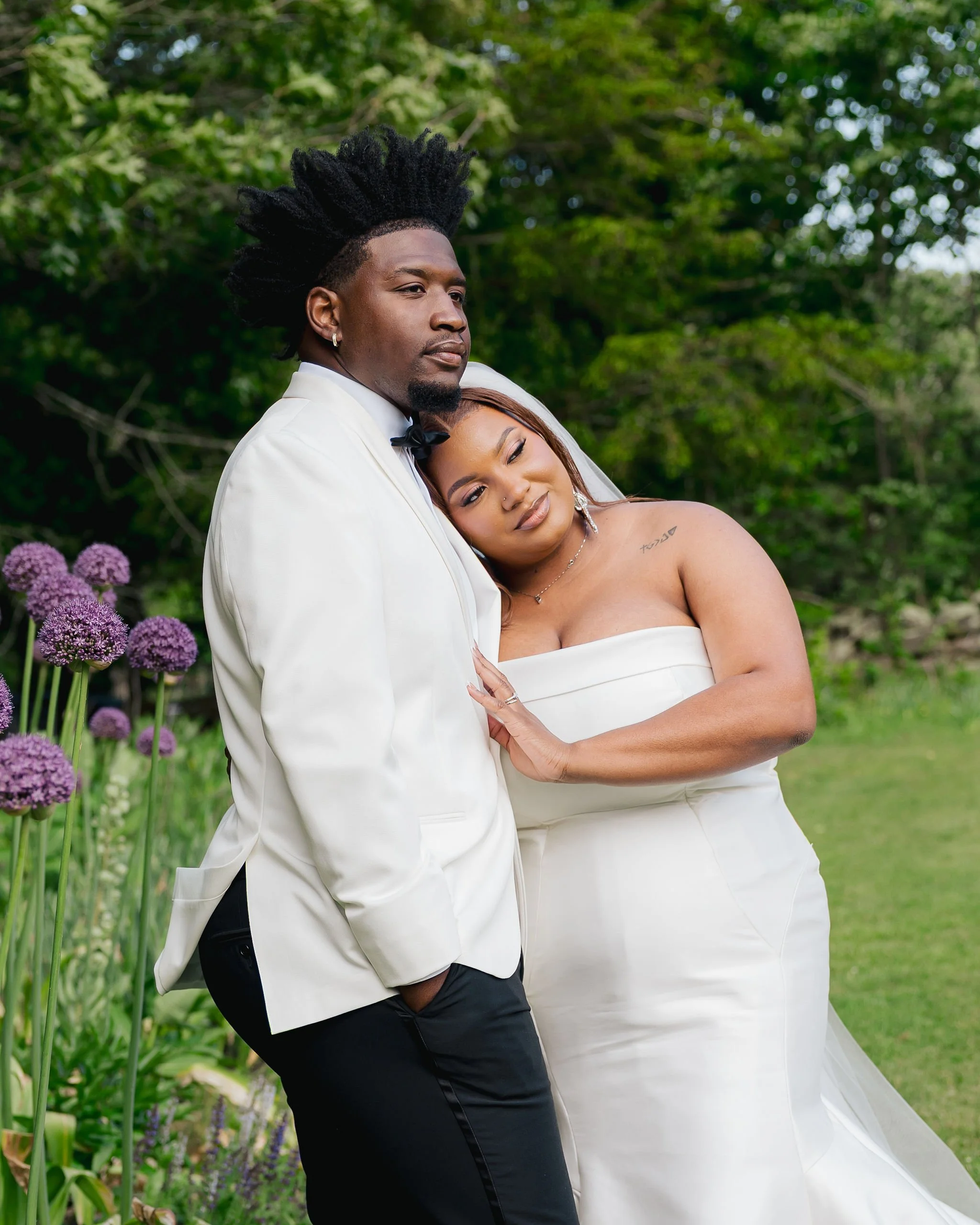 A couple dressed in wedding attire hugging outdoors with greenery and purple flowers in the background during a reception at Lambert's Cove Inn on Martha's Vineyard.