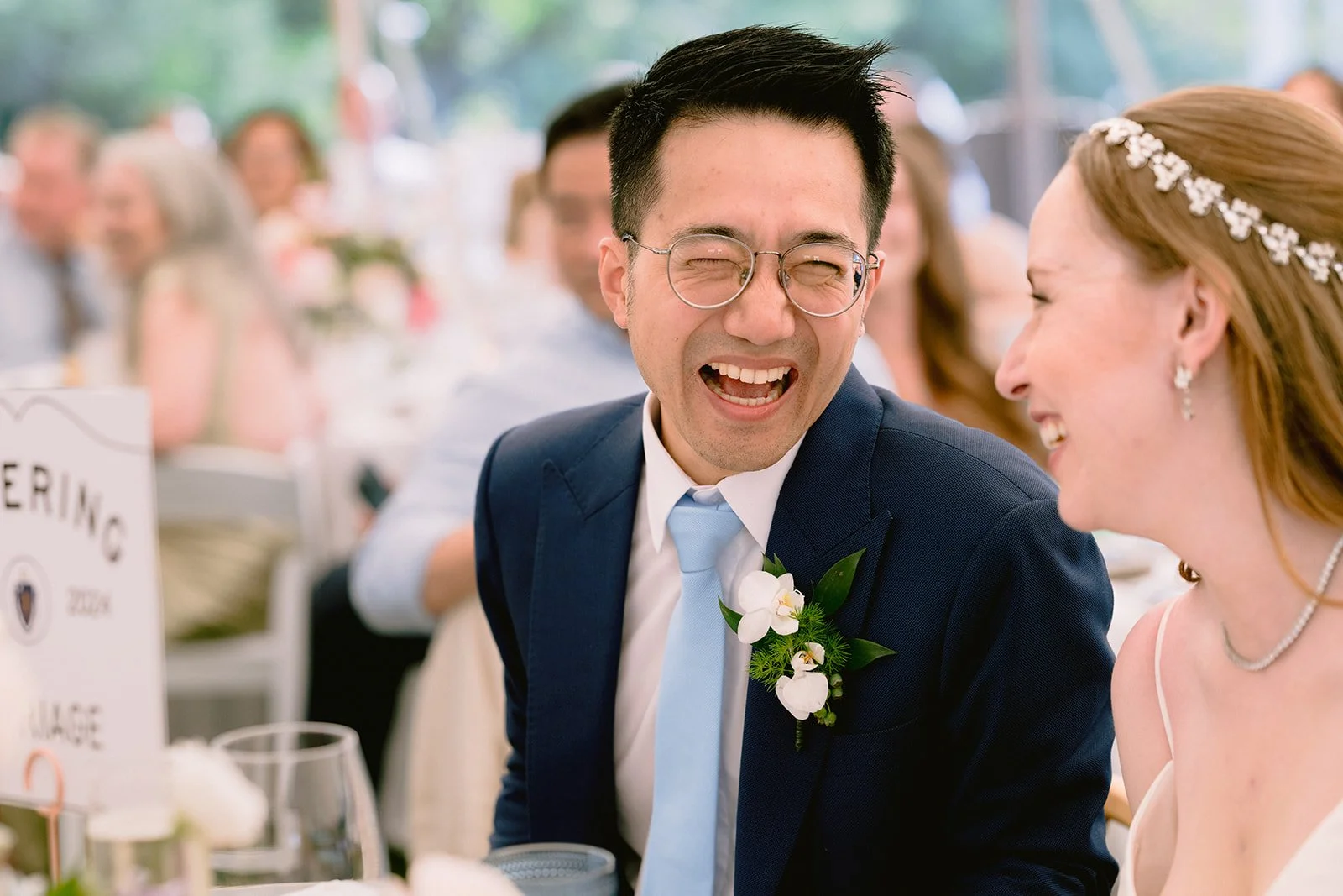 A man and woman laughing and smiling at a wedding reception. The man is wearing glasses, a navy blue suit, and a light blue tie with a white flower boutonniere. The woman is wearing a white dress and a floral headband. They are sitting at a decorated