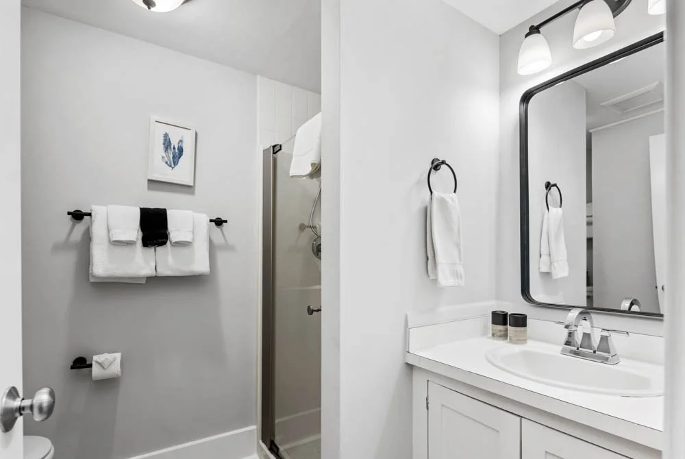 A clean, modern, white bathroom at Lambert's Cove Inn on Martha's Vineyard with a mirror, towel racks holding white towels, a shower stall, and decorative items on the sink counter.