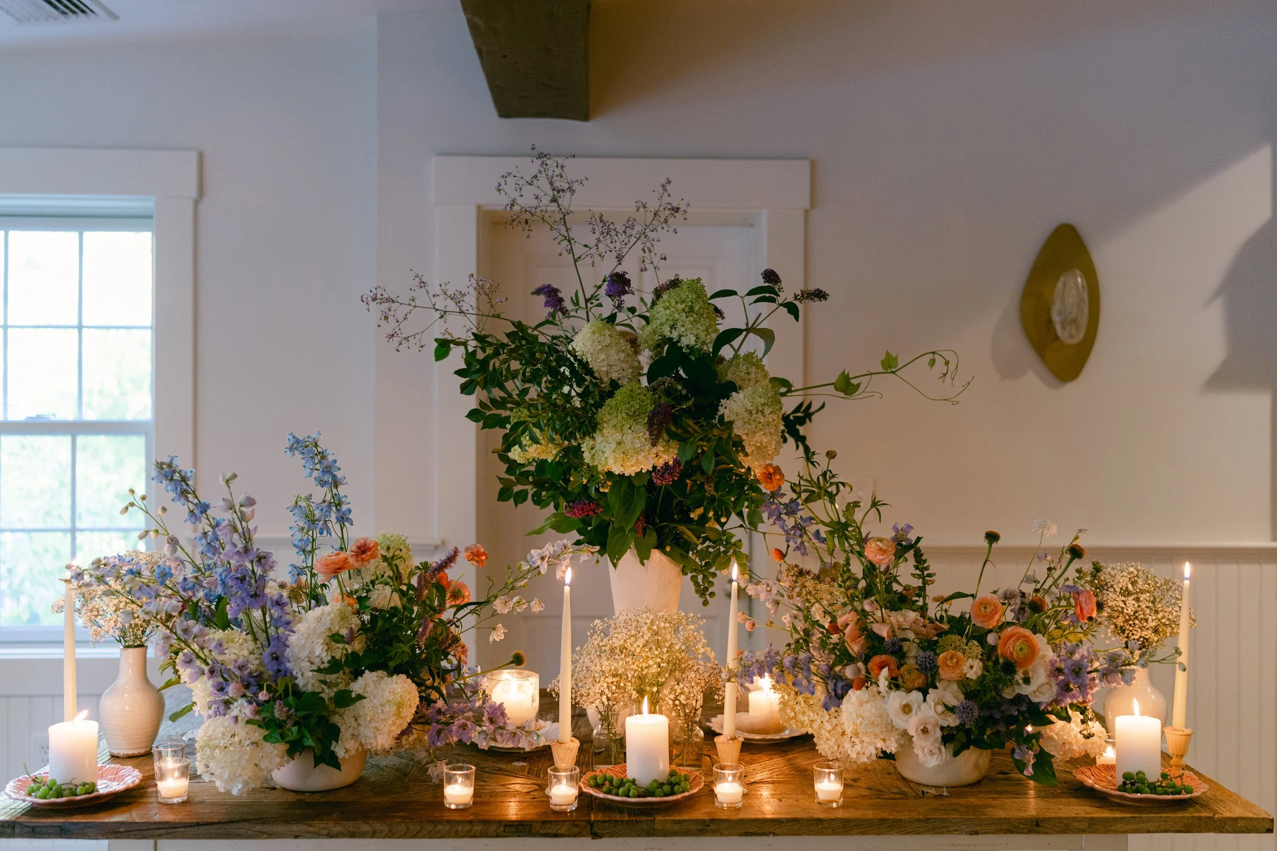 A centerpiece of various flowers including hydrangeas and roses, surrounded by candles and greenery, on a wooden table in a bright room with windows and wall decorations at Lambert's Cove Inn on Martha's Vineyard.