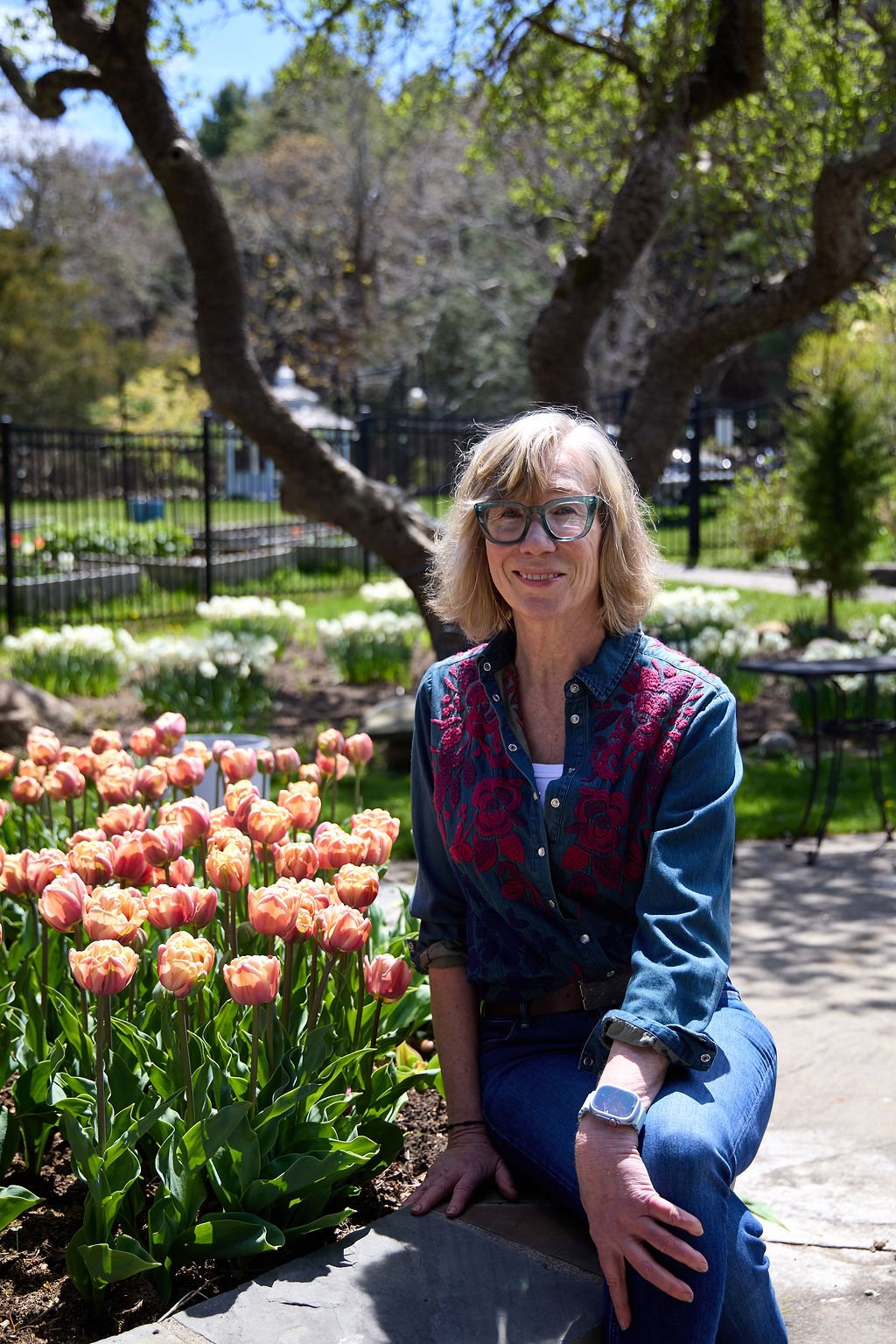 A woman with glasses and blonde hair sitting next to a bed of pink tulips in a garden.