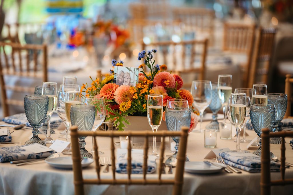 A round table set for a celebration with a centerpiece of pink, orange, and purple flowers, surrounded by champagne glasses and blue-patterned water glasses, in a decorated venue with wooden chairs.
