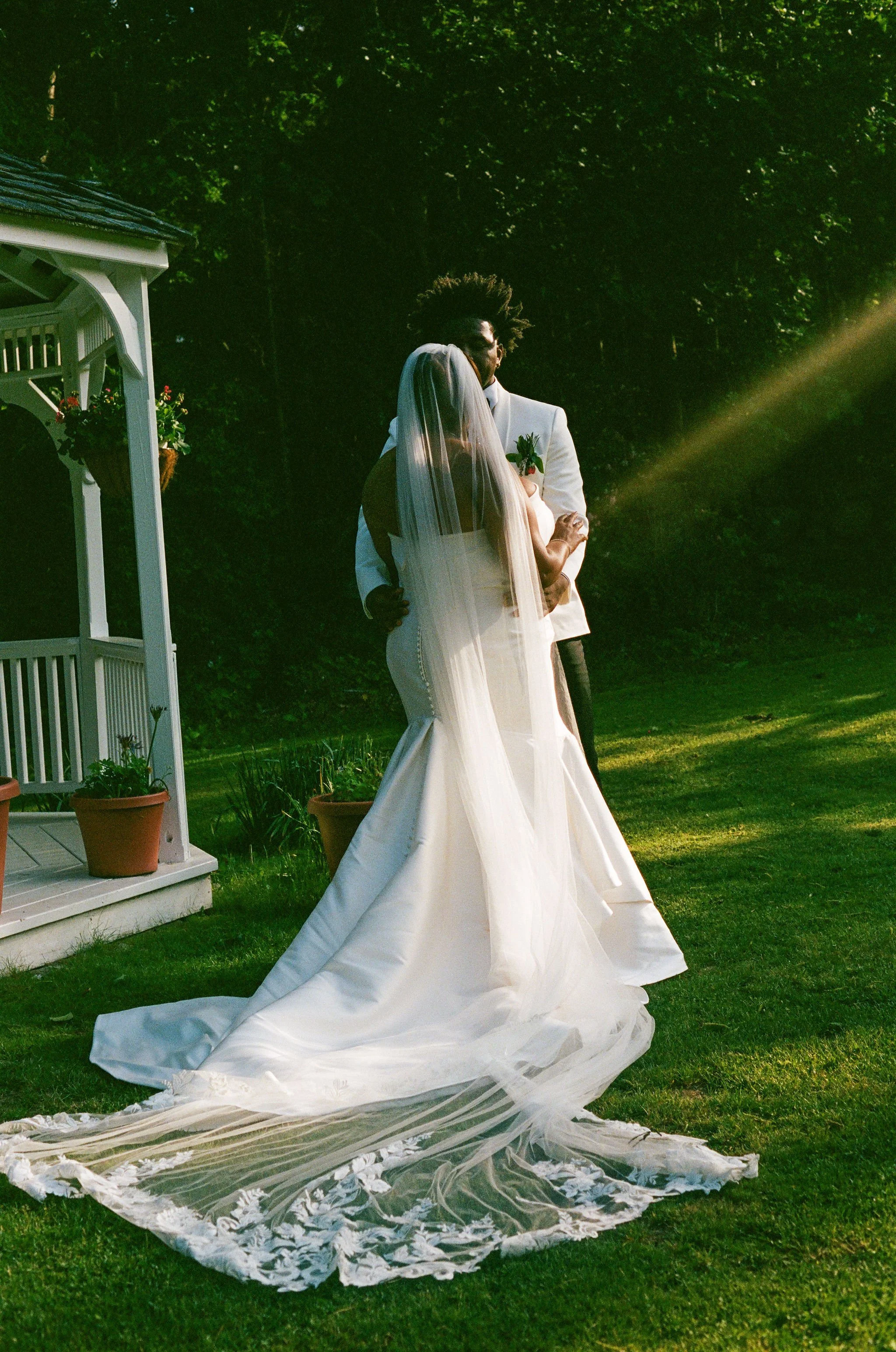 A bride and groom embracing outdoors during a wedding, with the bride wearing a long white dress and veil, and the groom in a white suit, standing on grass with trees in the background and a white garden gazebo nearby.