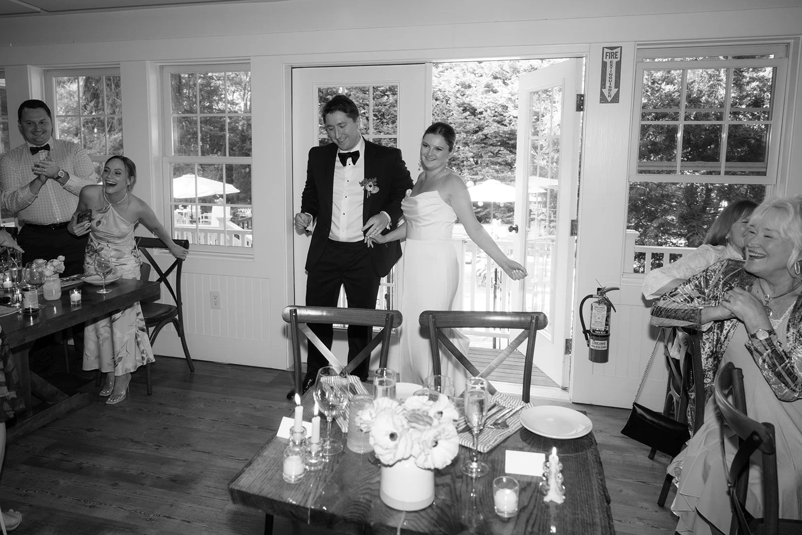 Black and white photo of a wedding celebration. Bride and groom are walking into the room; the bride is wearing a white dress, and the groom is in a tuxedo. Guests are sitting and standing around, smiling and laughing, with some holding drinks. The r
