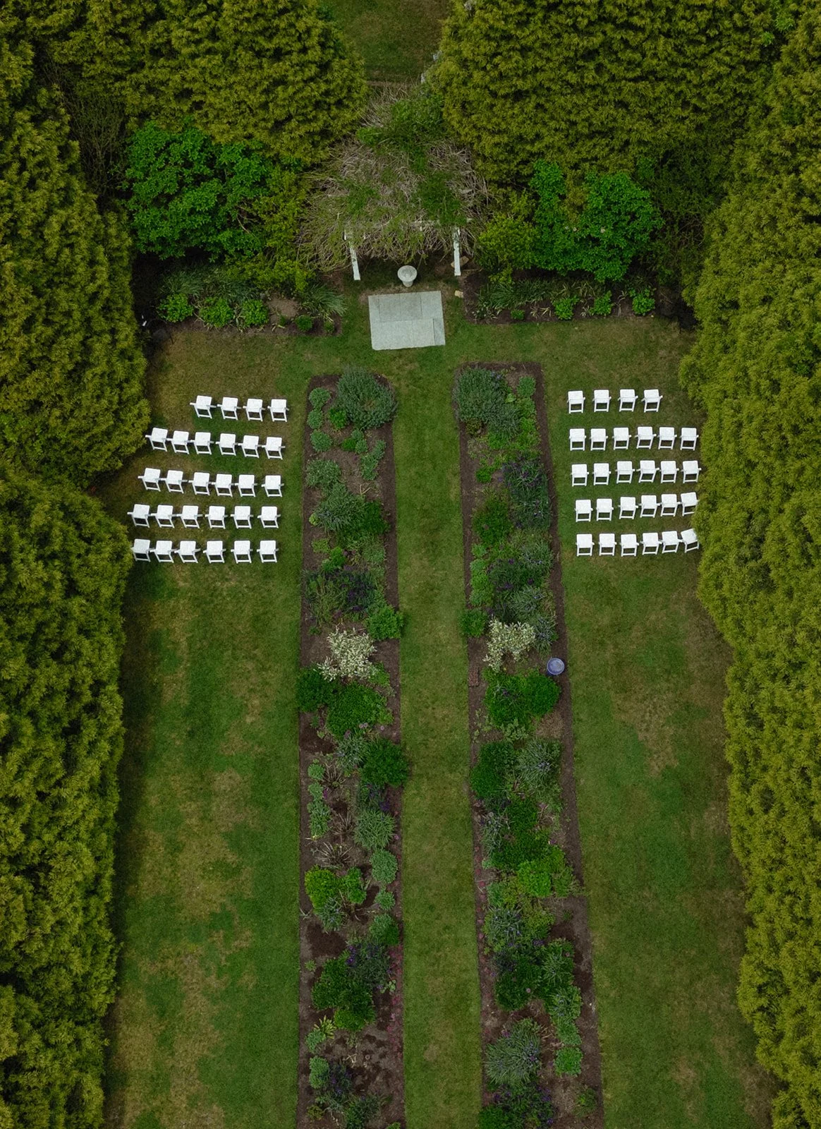 Aerial view of the outdoor wedding venue at Lambert's Cove Inn on Martha's Vineyard with flower beds, green plants, and a small thatched-roof shelter in the background. Rows of white chairs are arranged on either side of a central grassy walkway.