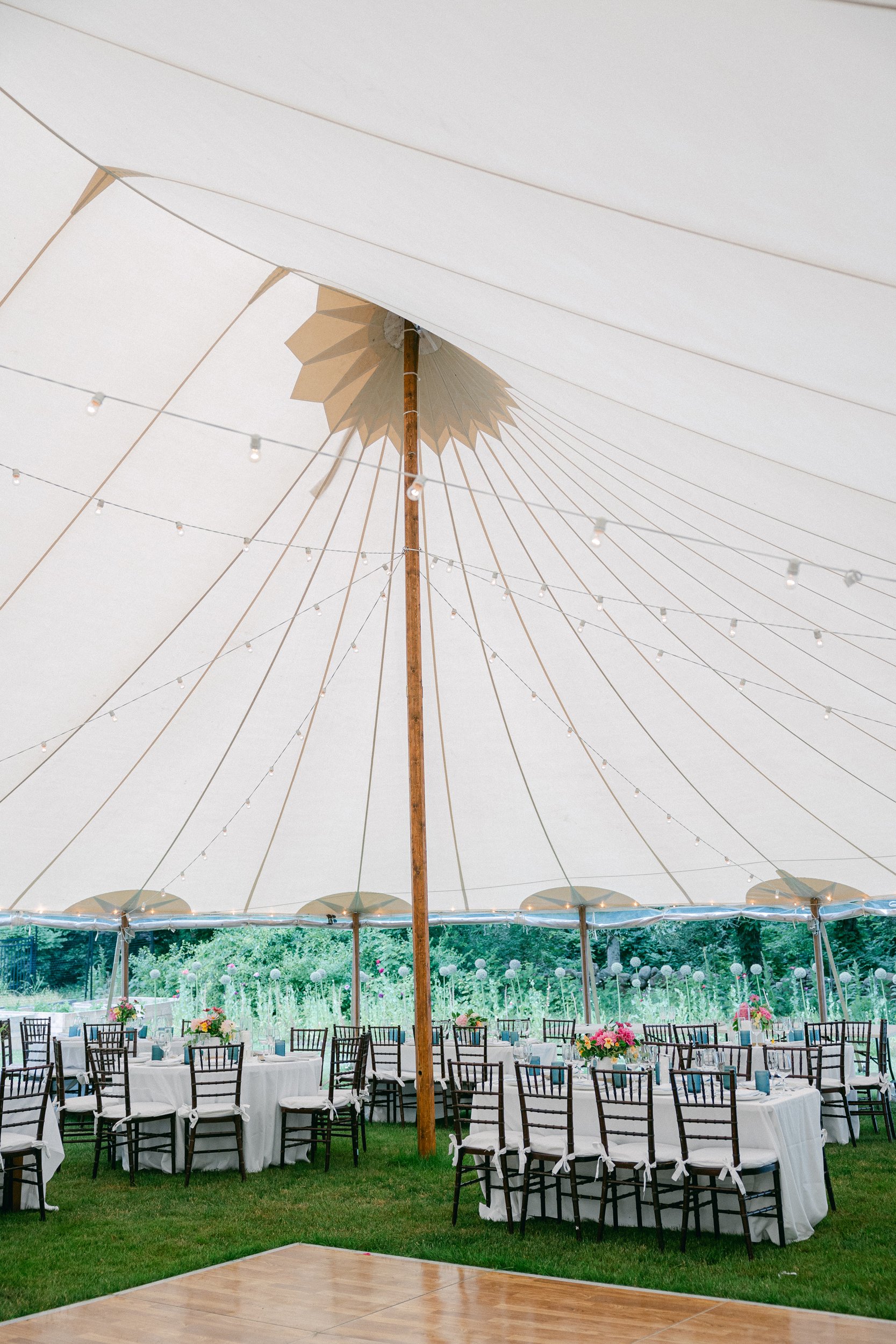 Event setup with round tables covered with white tablecloths, black chairs, floral centerpieces, under a large white canopy decorated with string lights and umbrellas, overlooking a landscaped garden during a wedding at Lambert's Cove Inn.