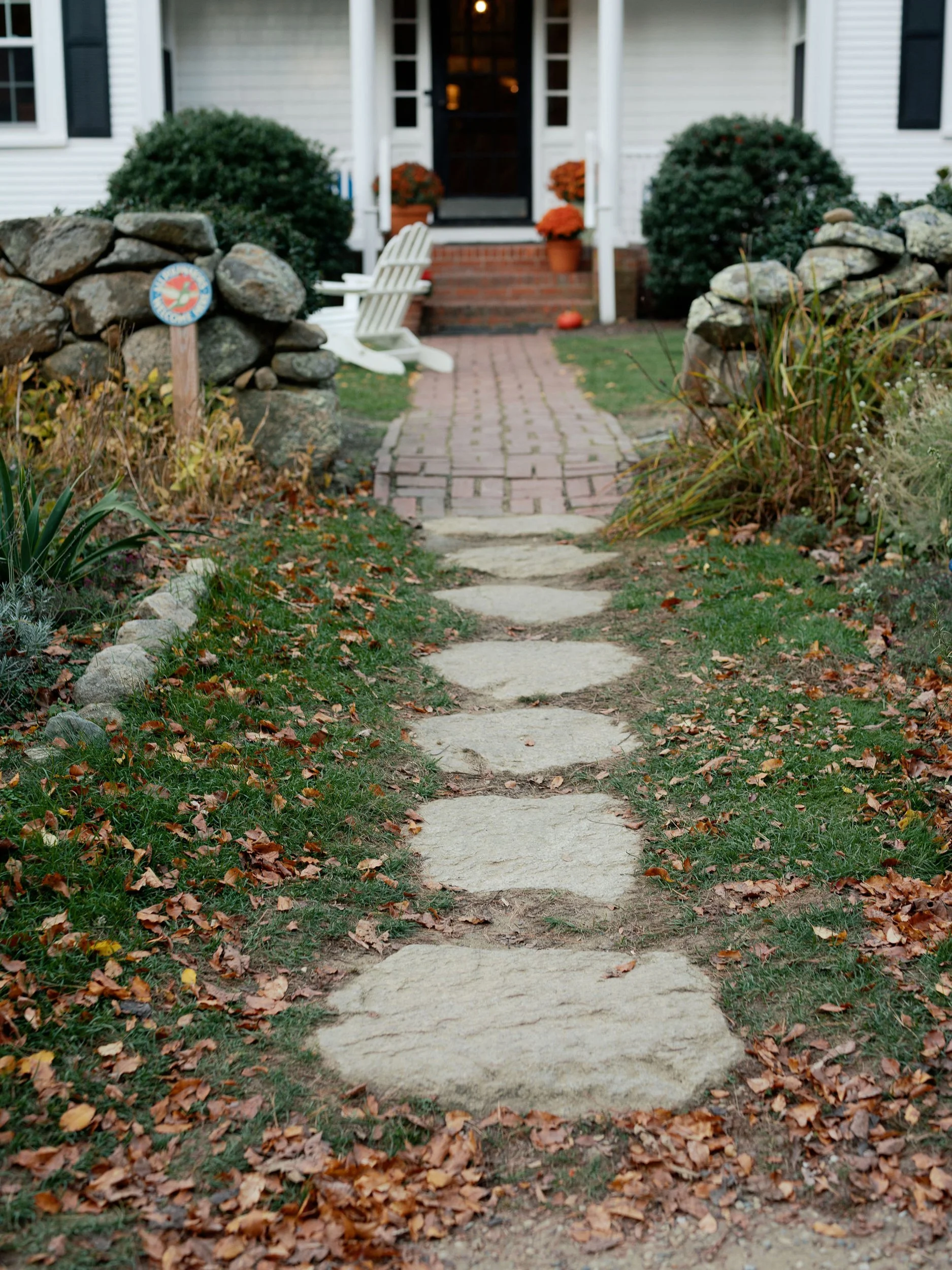 Stone pathway leading to a porch with brick steps, white chair, and orange pumpkins in front of a white house with black shutters, autumn leaves scattered on the grass.