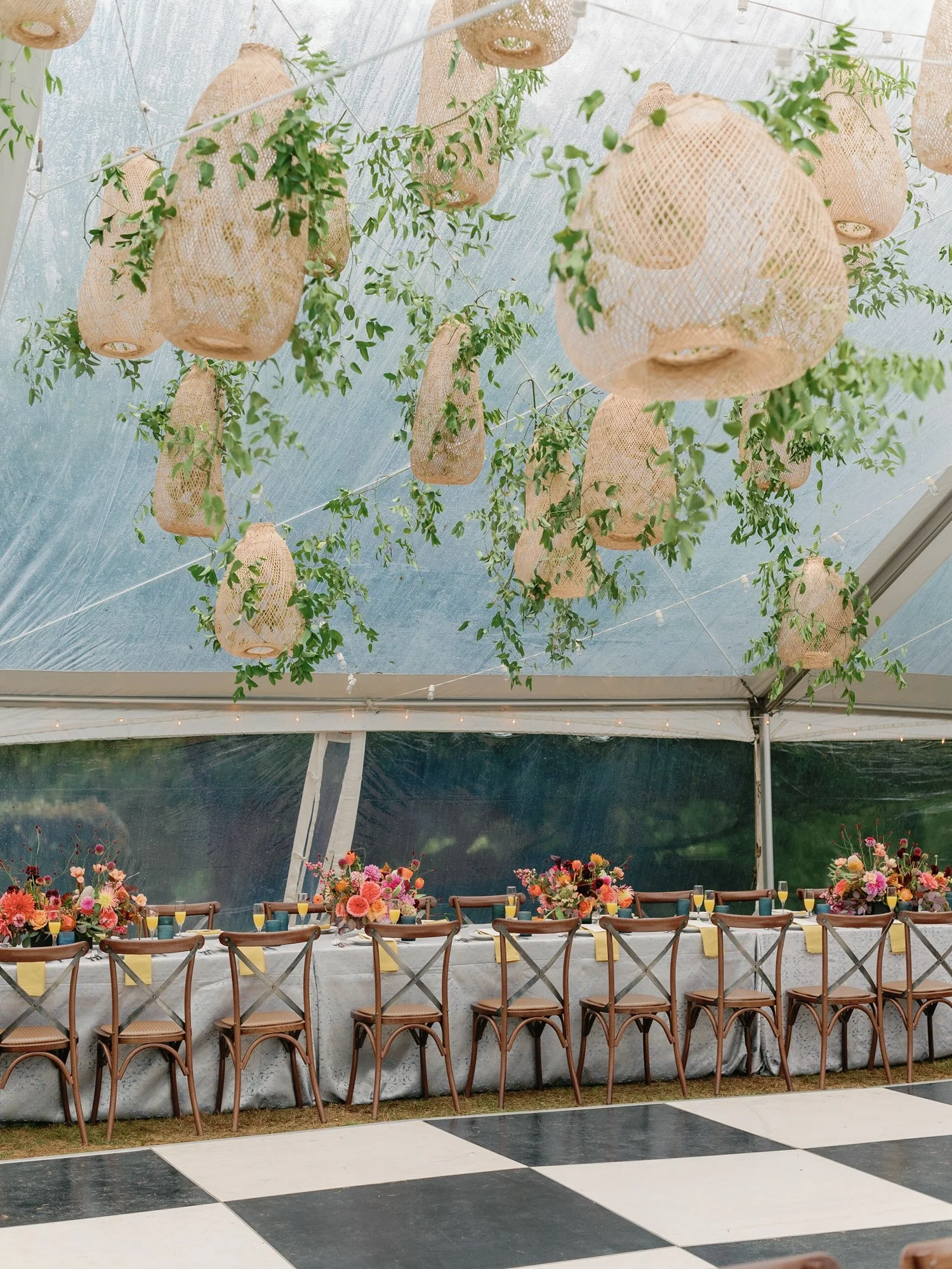A long rectangular table set for a celebration, decorated with colorful flower arrangements, surrounded by wooden chairs, under hanging woven lanterns and greenery in a tented event space at Lambert's Cove Inn on Martha's Vineyard.