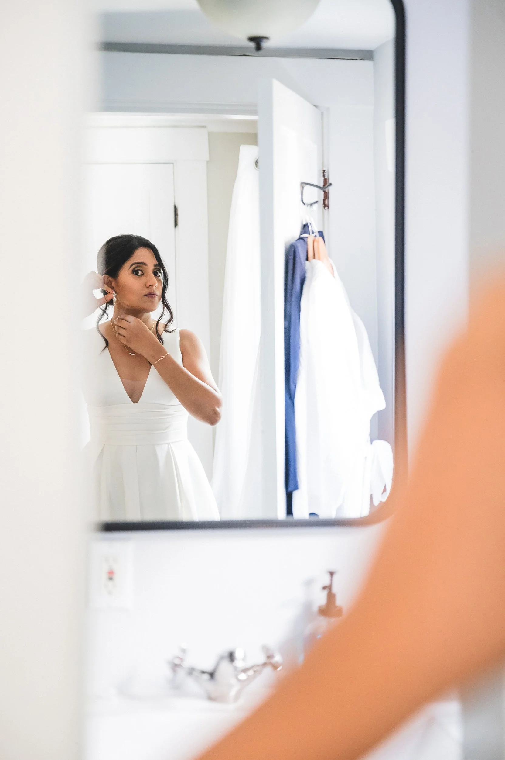 A woman in a white dress is looking at herself in a mirror, adjusting her earring, in a room with white walls at Lambert's Cove Inn on Martha's Vineyard. There are clothes hanging on the wall and a sink with a soap dispenser in the foreground.