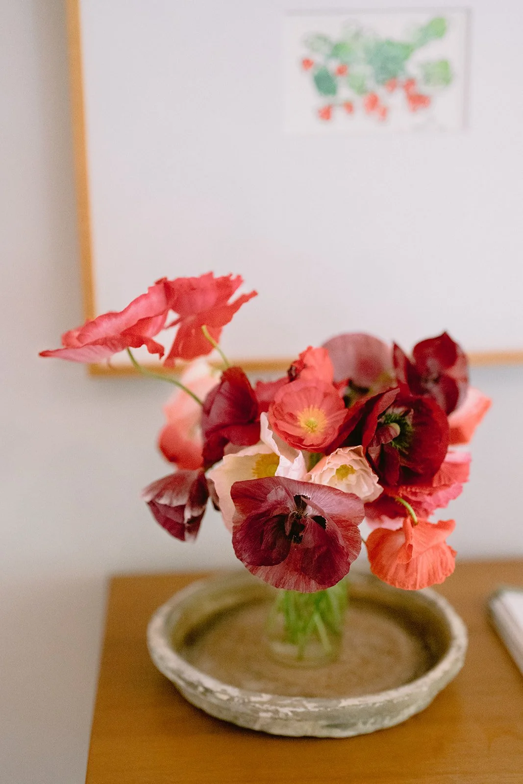 A bouquet of pink, red, and white flowers in a small, round, textured vase on a wooden surface, with a whiteboard and colorful map of Europe in the background.