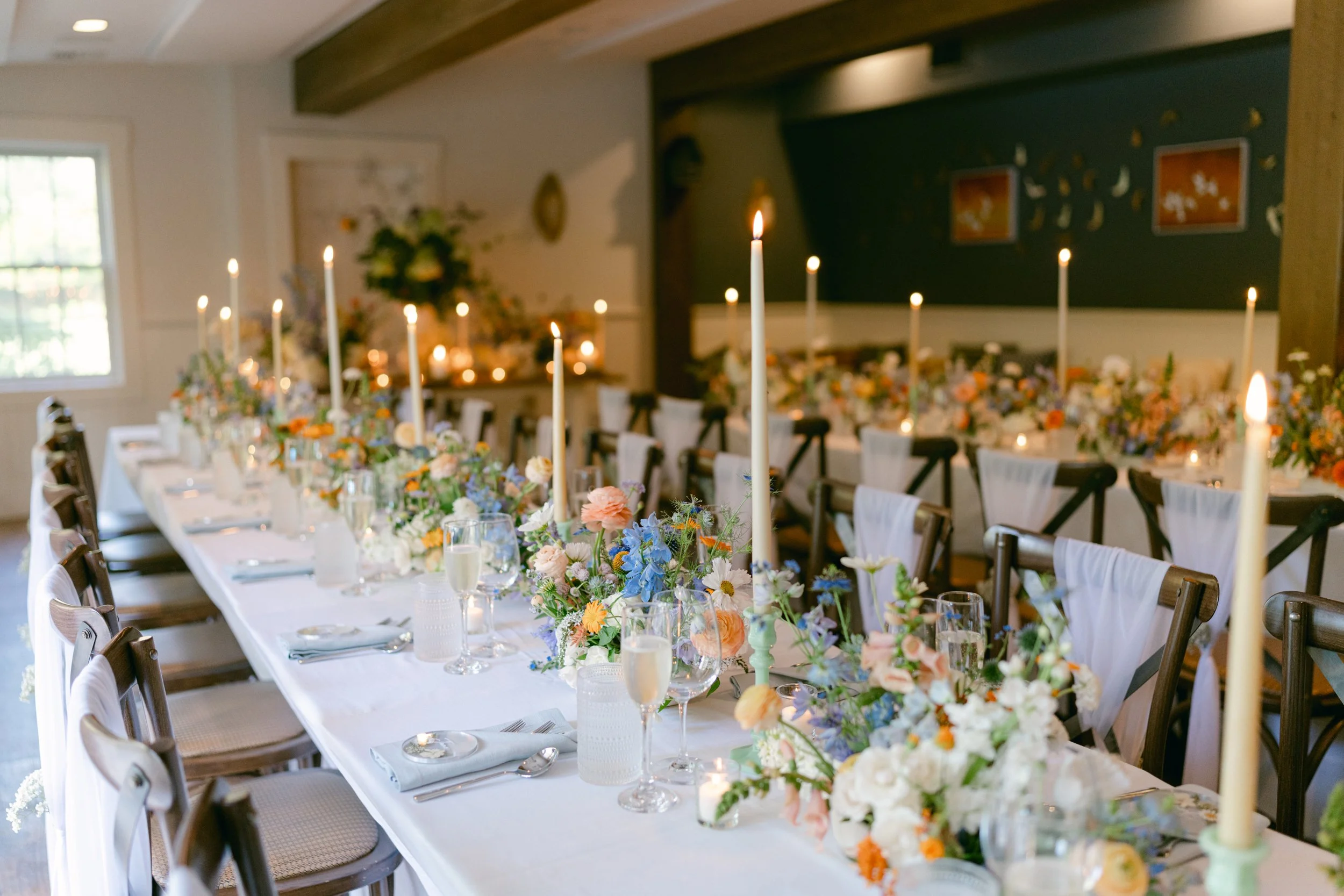 Elegant indoor dining table decorated with floral centerpieces and tall candleholders, set for a special occasion. The room has natural light, a window on the left, and artwork on the back wall at Lambert's Cove Inn on Martha's Vineyard.