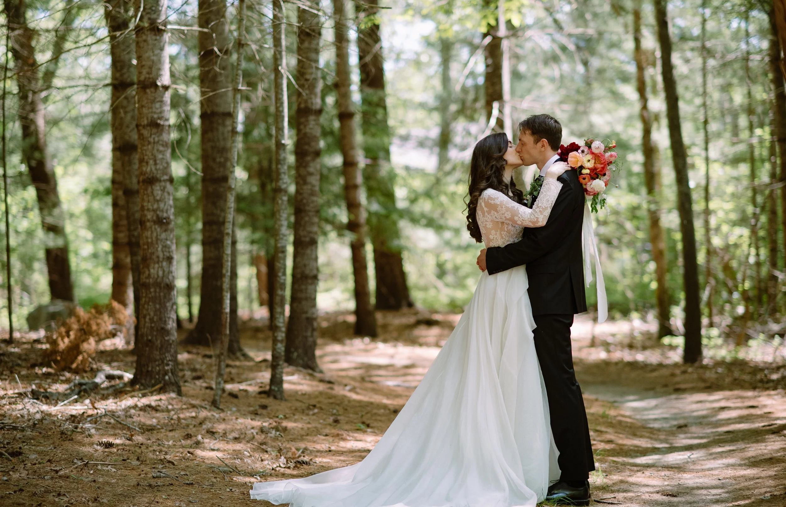 A bride and groom kiss surrounded by trees during a wedding at Lambert's Cove Inn on Martha's Vineyard. The bride holds a bouquet of red, pink, and white flowers, and the bride wears a lace wedding dress, while the groom wears a black suit.