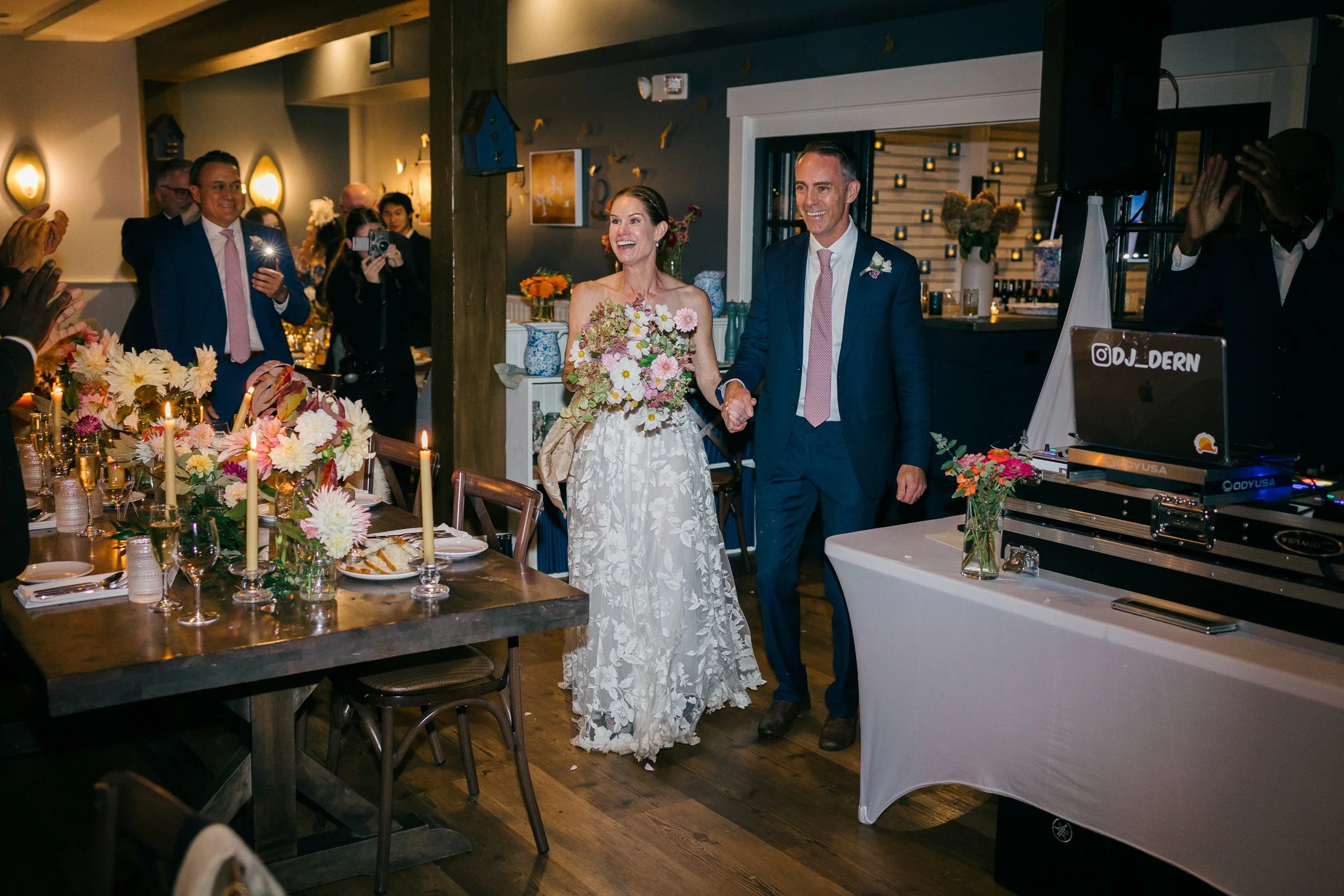 A bride and groom holding hands, smiling, entering a reception at Lambert's Cove Inn on Martha's Vineyard decorated with flowers and candles. Guests are taking photos and celebrating.