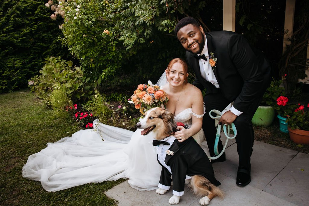A bride and groom are posing with a dog dressed in a tuxedo, outside in a garden with flowers and greenery during a wedding reception at Lambert's Cove Inn on Martha's Vineyard.