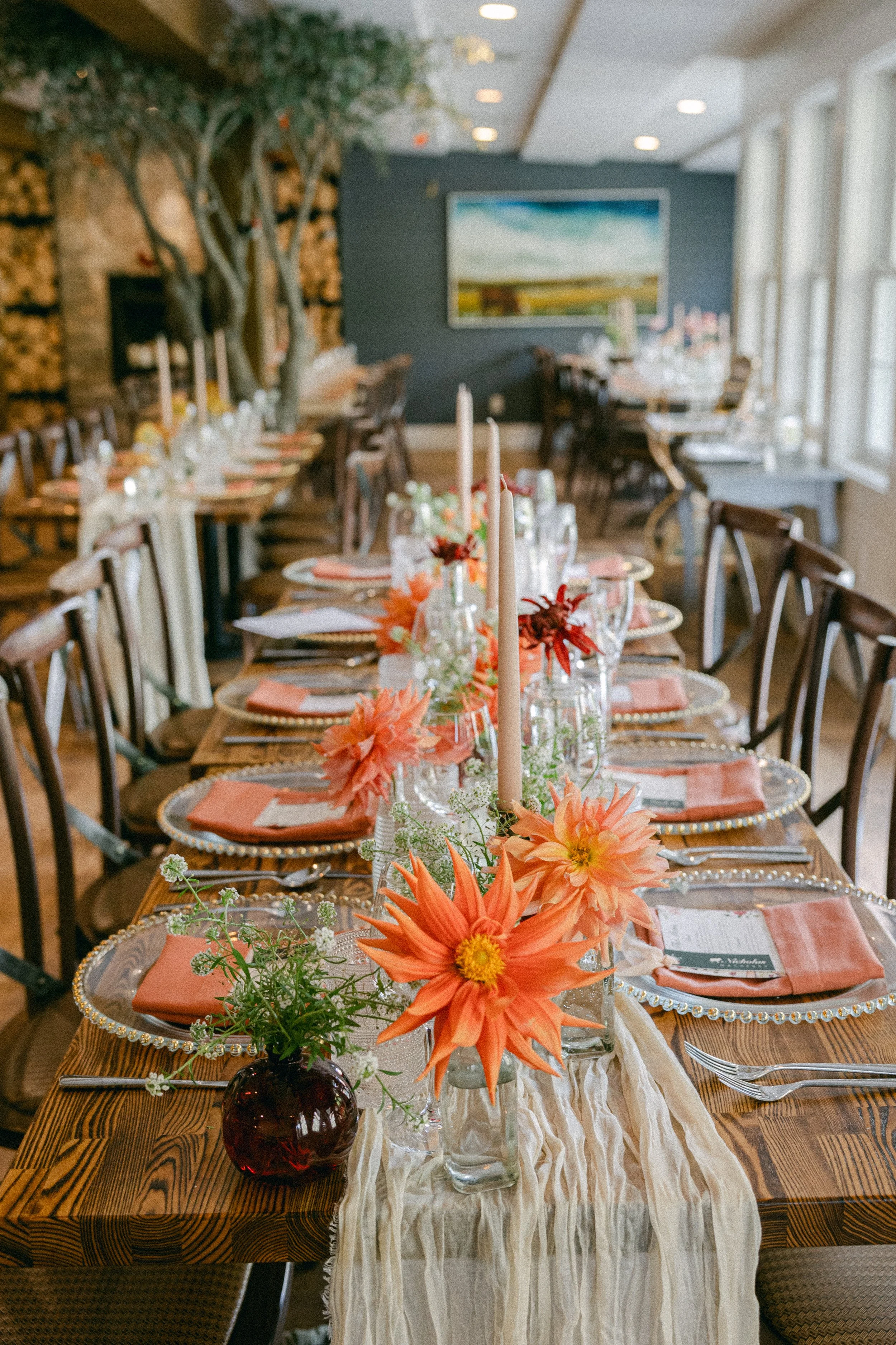 A long wooden dining table set for a special occasion with pink napkins, clear glasses, silverware, and floral centerpieces featuring orange flowers in a dark vase and smaller white flowers. The table is covered with a light-colored table runner, and