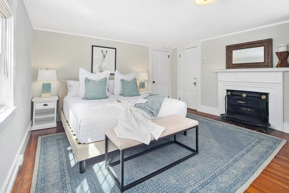 A cozy bedroom with white walls, a white bed with light green pillows, and two bedside lamps. There is a fireplace with a mirror and decorative items above it, and a blue area rug on the wooden floor.