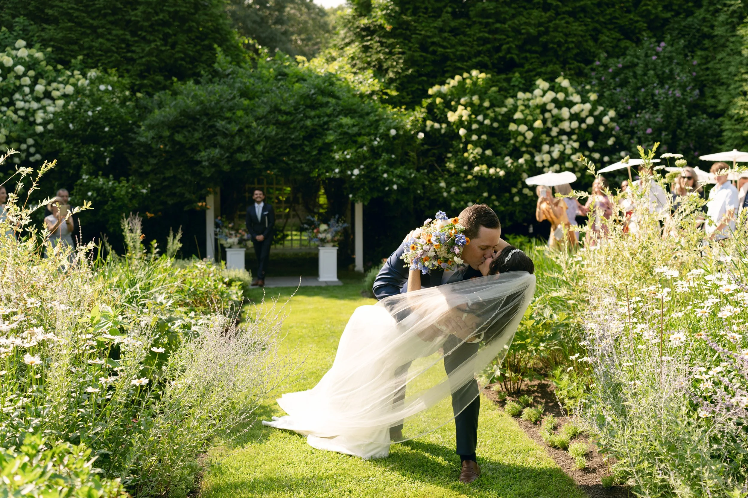 A couple kissing outdoors during a wedding ceremony in the English garden at Lambert's Cove Inn on Martha's Vineyard, surrounded by custom grown flowers, with guests in the background.