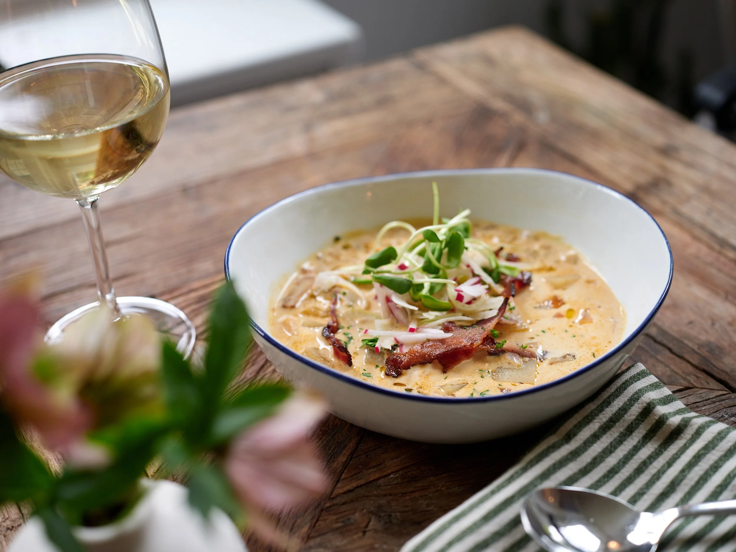 A bowl of creamy soup garnished with microgreens and bacon, with a glass of white wine and a striped cloth on a wooden dining table.