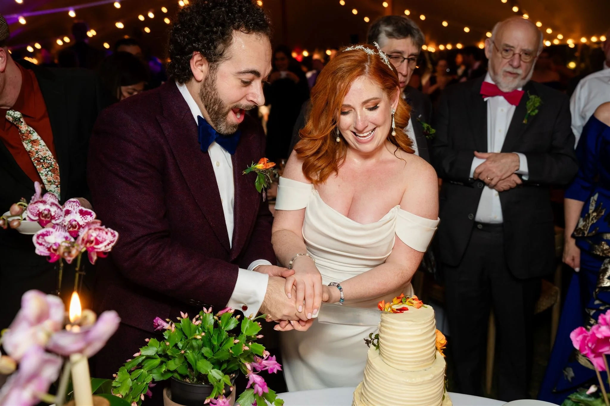 A bride and groom cut a wedding cake together at their wedding reception, surrounded by guests and floral decorations at Lambert's Cove Inn on Martha's Vineyard.