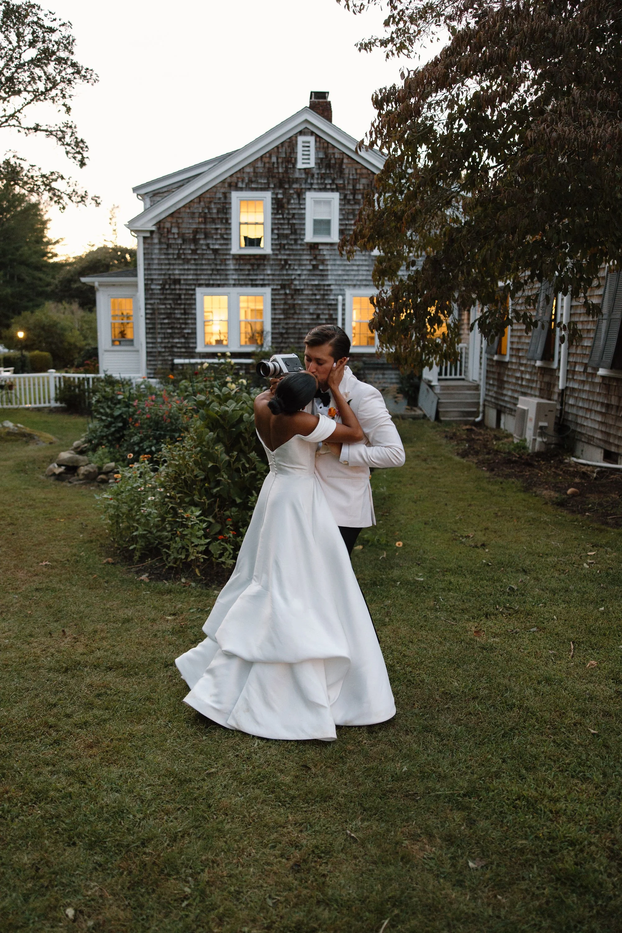 A bride and groom share a kiss while dancing outside in a garden near at Lambert's Cove Inn on Martha's Vineyard with illuminated windows at dusk.