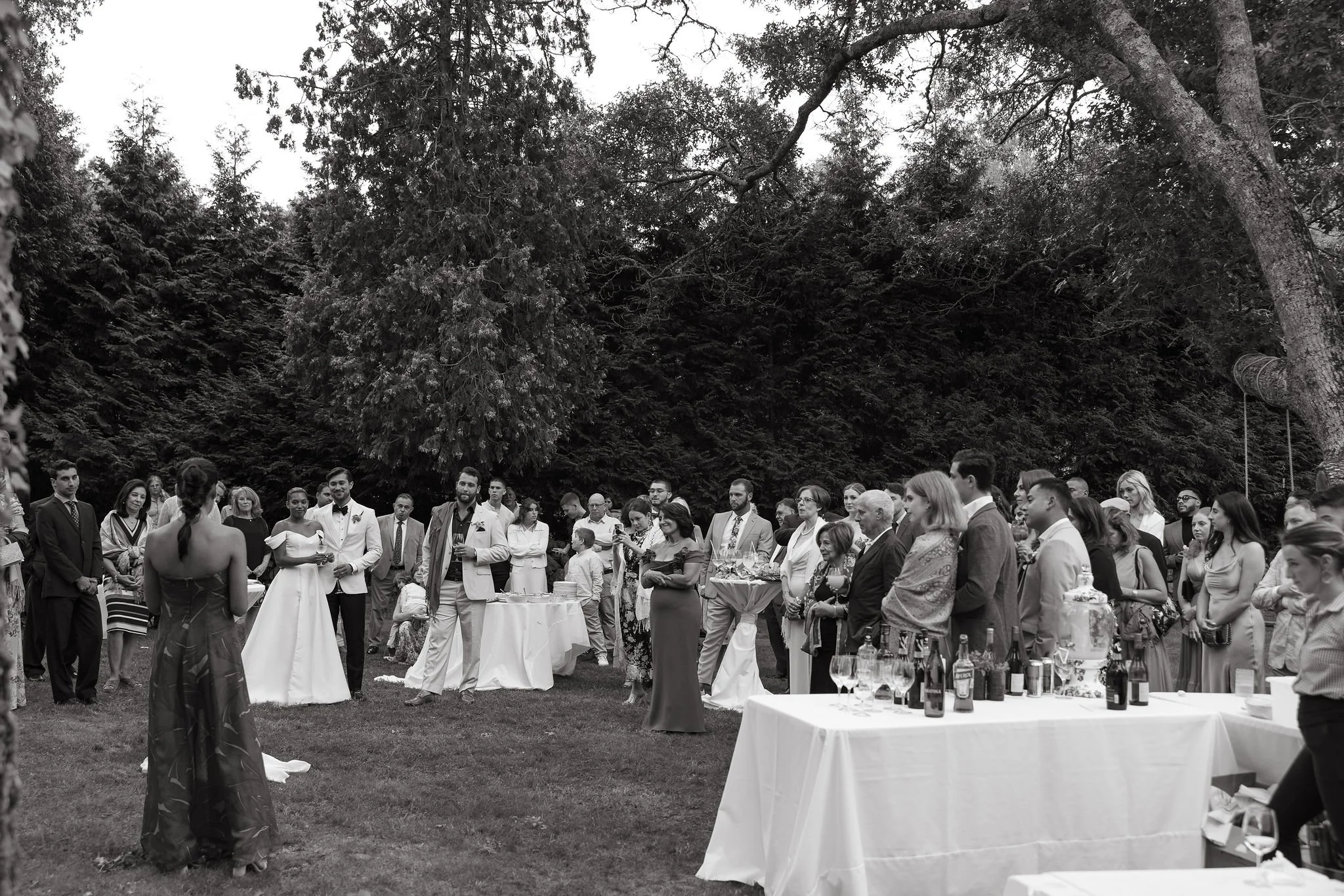 A large gathering of people outdoors for an outdoor wedding reception at Lambert's Cove Inn on Martha's Vineyard, with tables of drinks and a mountainous or forested background.
