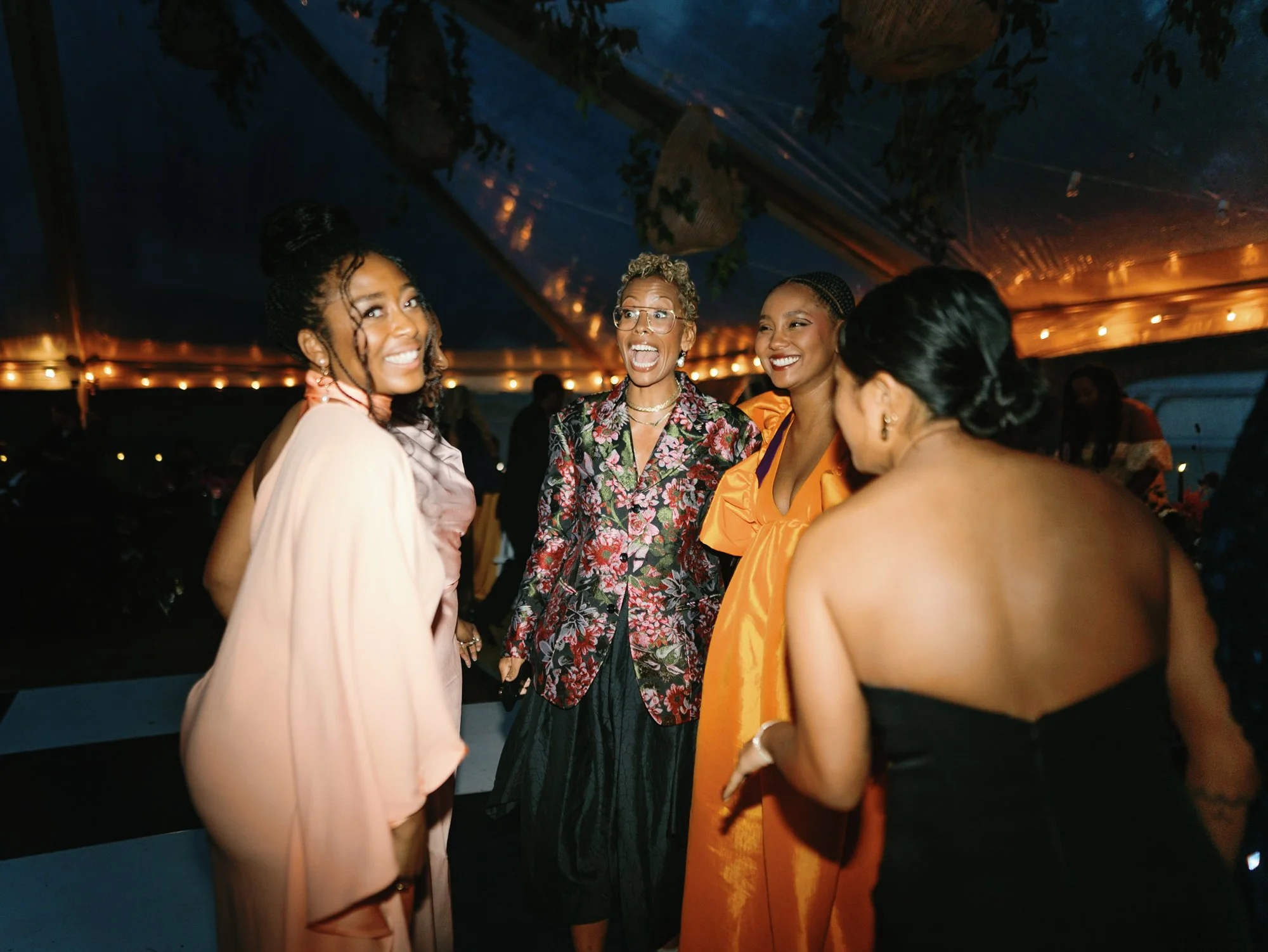 A group of five women smiling and talking at a wedding reception at Lambert's Cove Inn on Martha's Vineyard with warm lighting, dressed in colorful and elegant clothing.