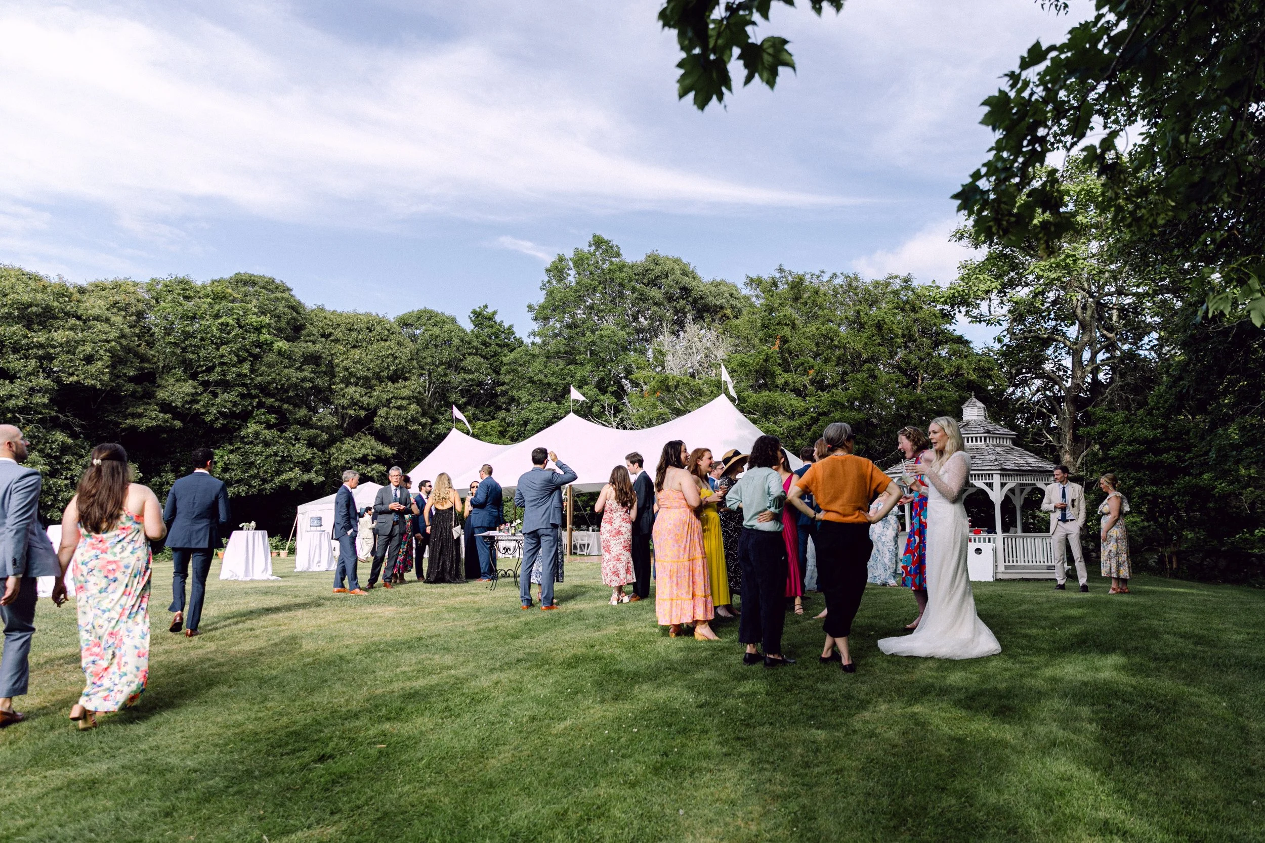 People mingling outdoors at a wedding reception at Lambert's Cove Inn on Martha's Vineyard in West Tisbury with a white tent, surrounded by trees and a gazebo in the background.