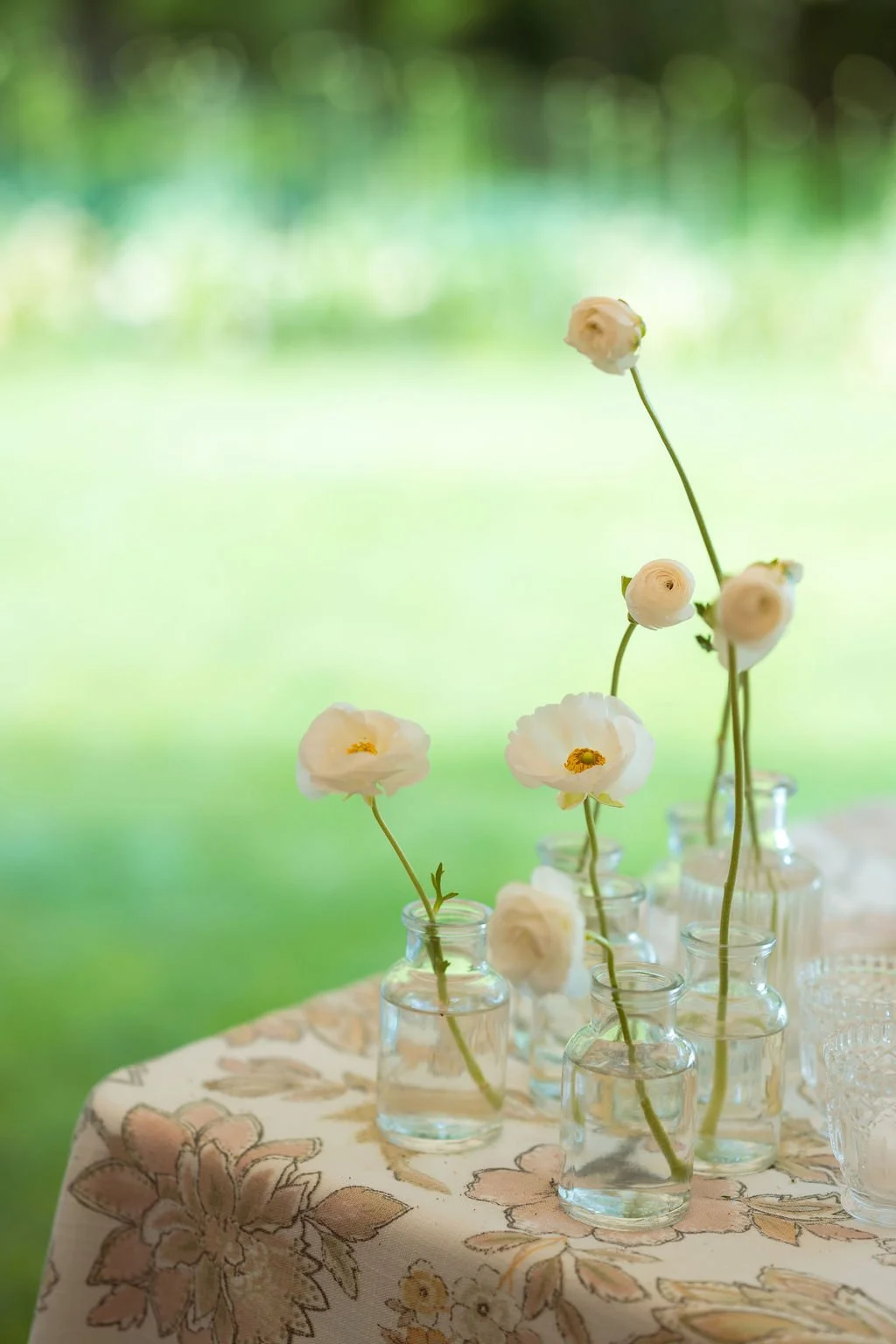 Seven small glass bottles with water, each holding a delicate white flower with yellow centers, arranged on a table with a floral patterned tablecloth against a blurred green outdoor background at Lambert's Cove Inn on Martha's Vineyard.