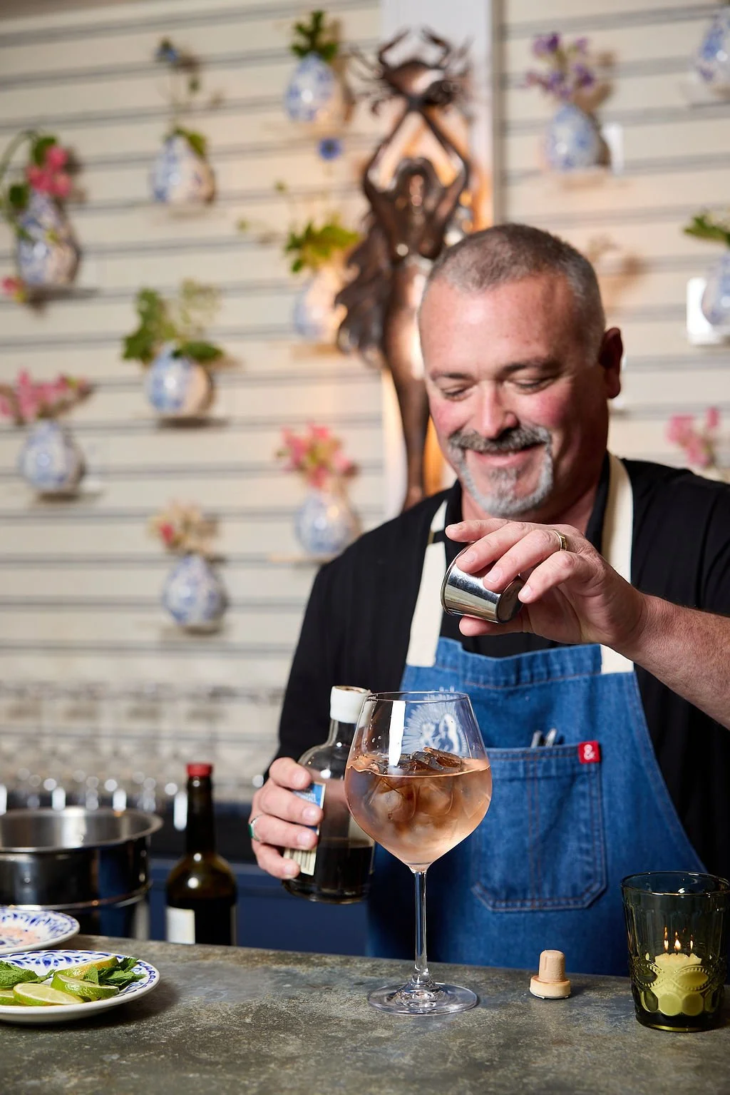 A smiling man in a black shirt and blue apron is pouring a dark liquid into a wine glass filled with ice at Mermaid Bar at Lambert's Cove Inn on Martha's Vineyard.