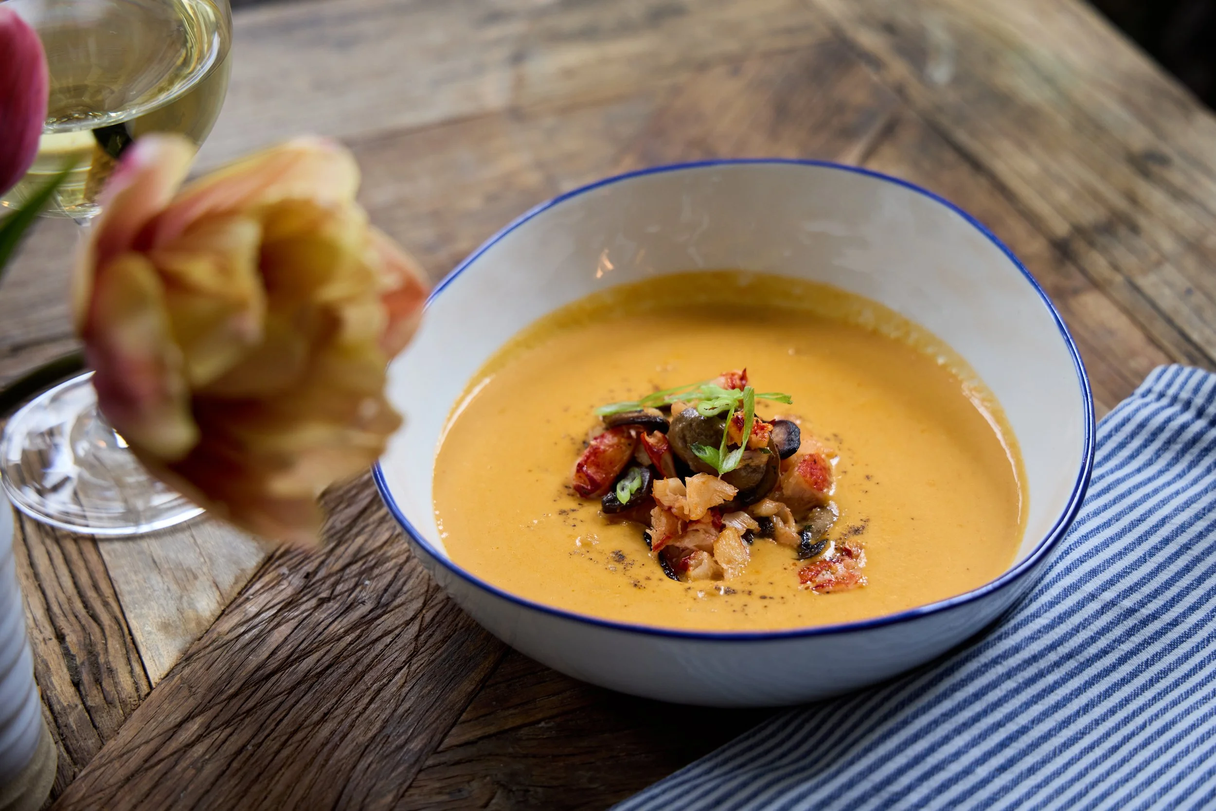 A bowl of creamy orange soup topped with chopped vegetables and herbs on a wooden table, next to a glass of white wine and an artichoke at Restaurant Foster at Lambert's Cove Inn on Martha's Vineyard.