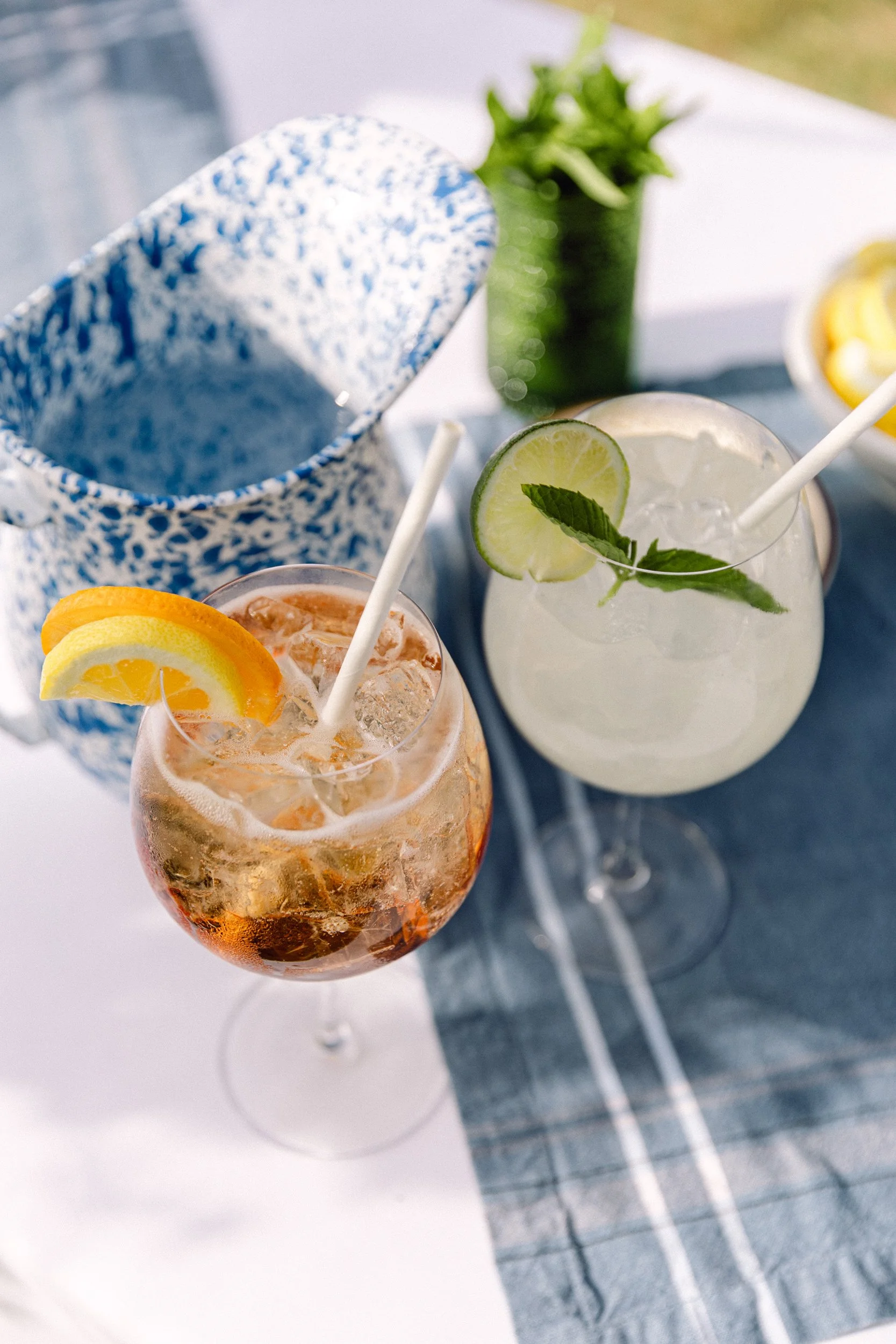 Two colorful cocktails with lemon and lime slices and mint leaves, on a table with a blue and white fabric, and a green plant in the background at Lambert's Cove Inn on Martha's Vineyard.