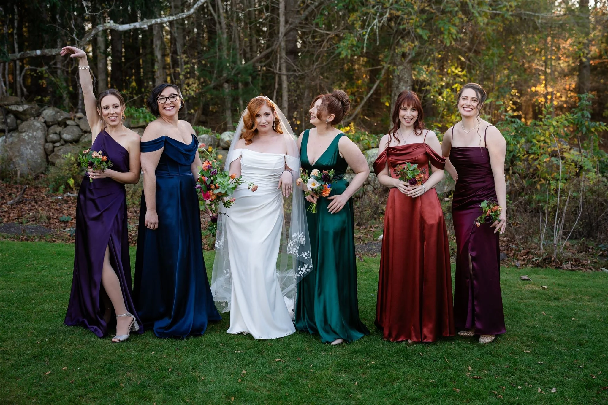 A group of six women, including a bride in a white wedding dress and five bridesmaids in colorful dresses, standing outdoors on green grass with a wooded background during sunset. They are smiling and holding bouquets.