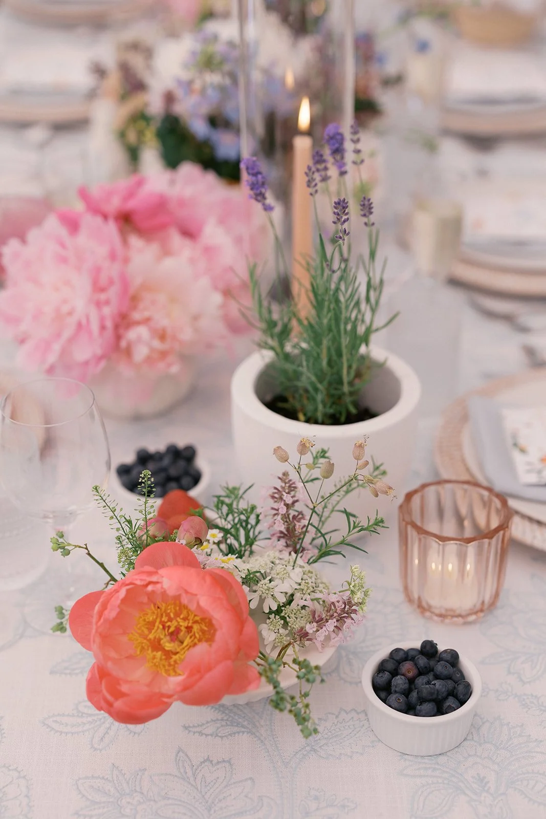 Table setting with pink, purple, and white flowers in vases, bowls of blueberries, candles, plates, and glassware on a white tablecloth.