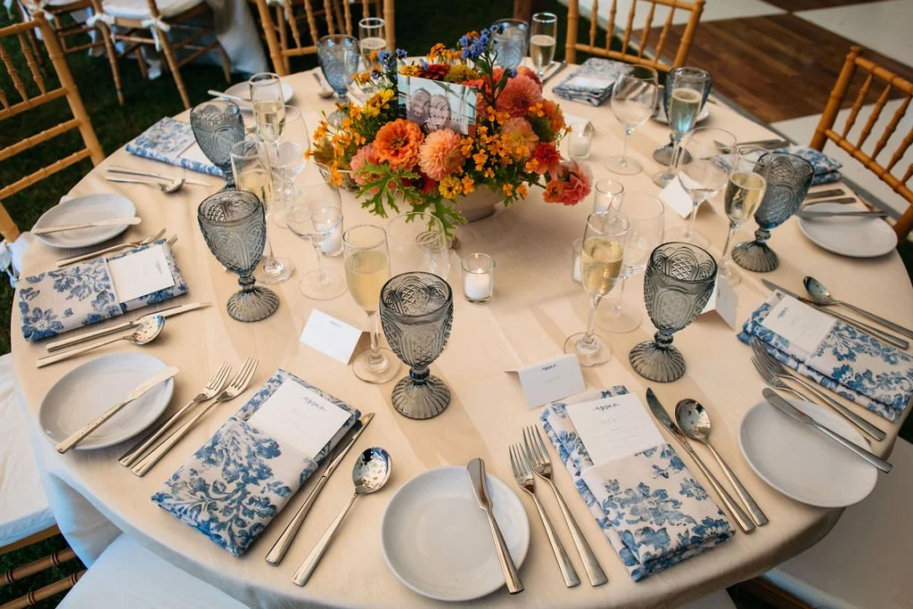 Round banquet table set with floral centerpiece, champagne glasses, water glasses, silverware, blue and white napkins, white plates, and black goblets, for a formal event.