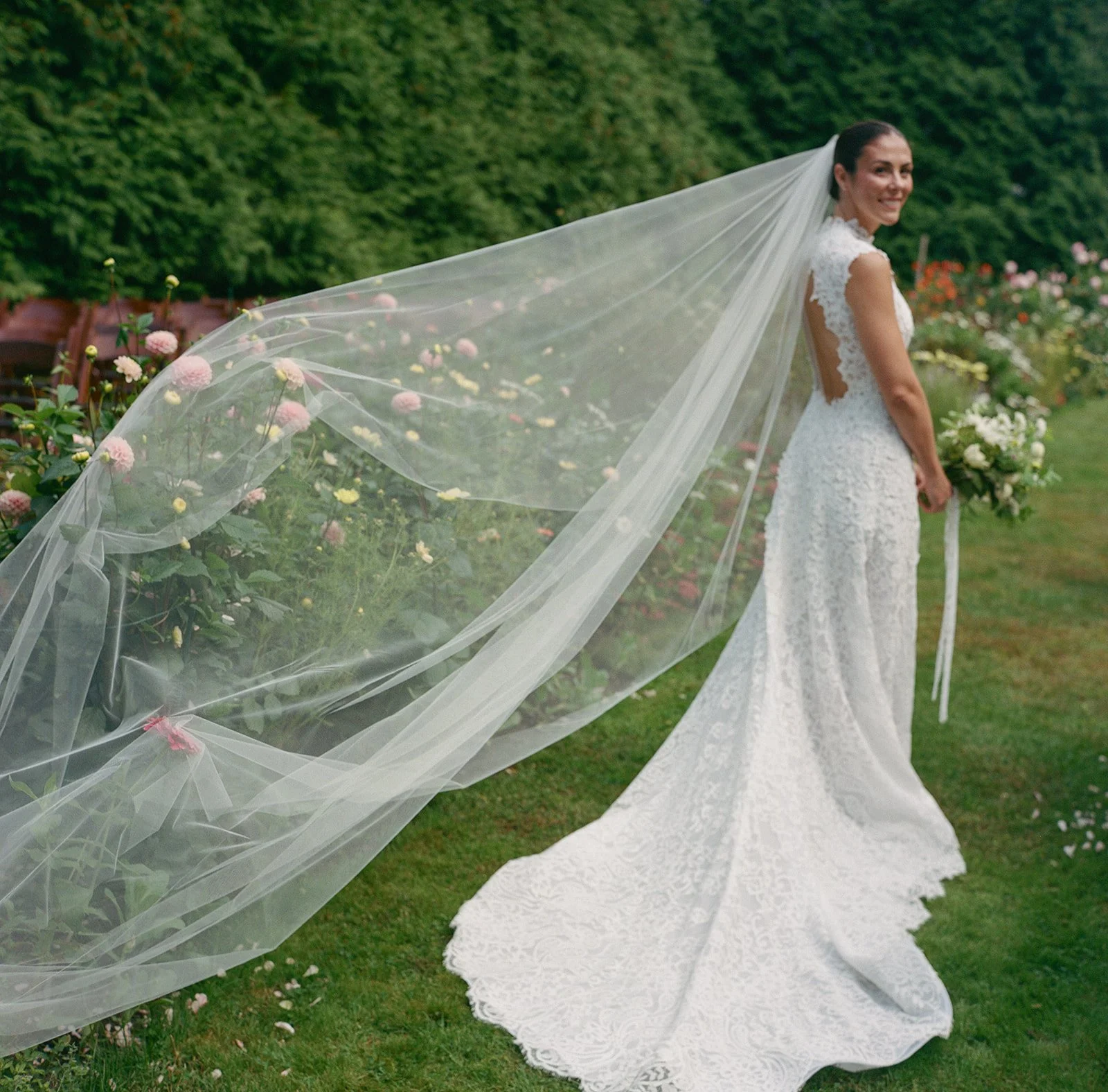 A smiling bride in a white lace wedding dress with a long train, standing outdoors in a garden with flowers and greenery, holding a bouquet, with a sheer veil flowing behind her during a wedding at Lambert's Cove Inn on Martha's Vineyard.