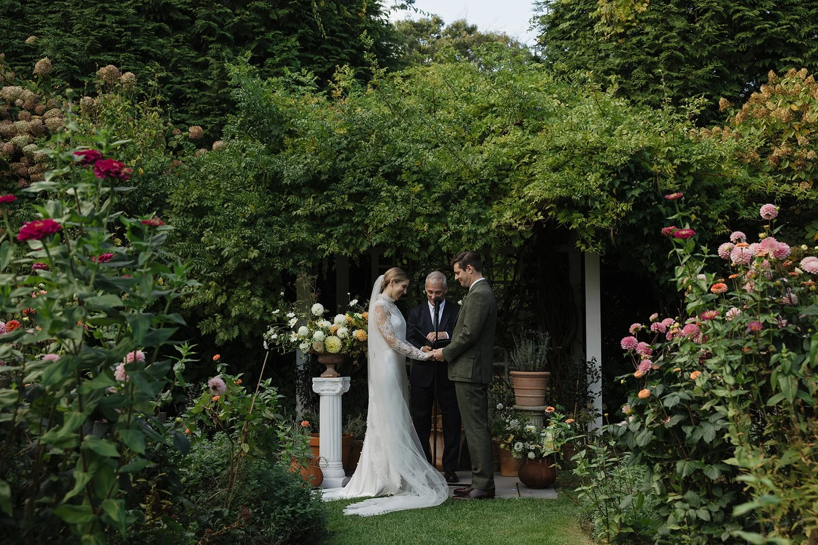 A bride and groom standing hand in hand during their outdoor wedding ceremony at Lambert's Cove Inn on Martha's Vineyard, with an officiant reading vows. They are surrounded by lush greenery and colorful flowers.
