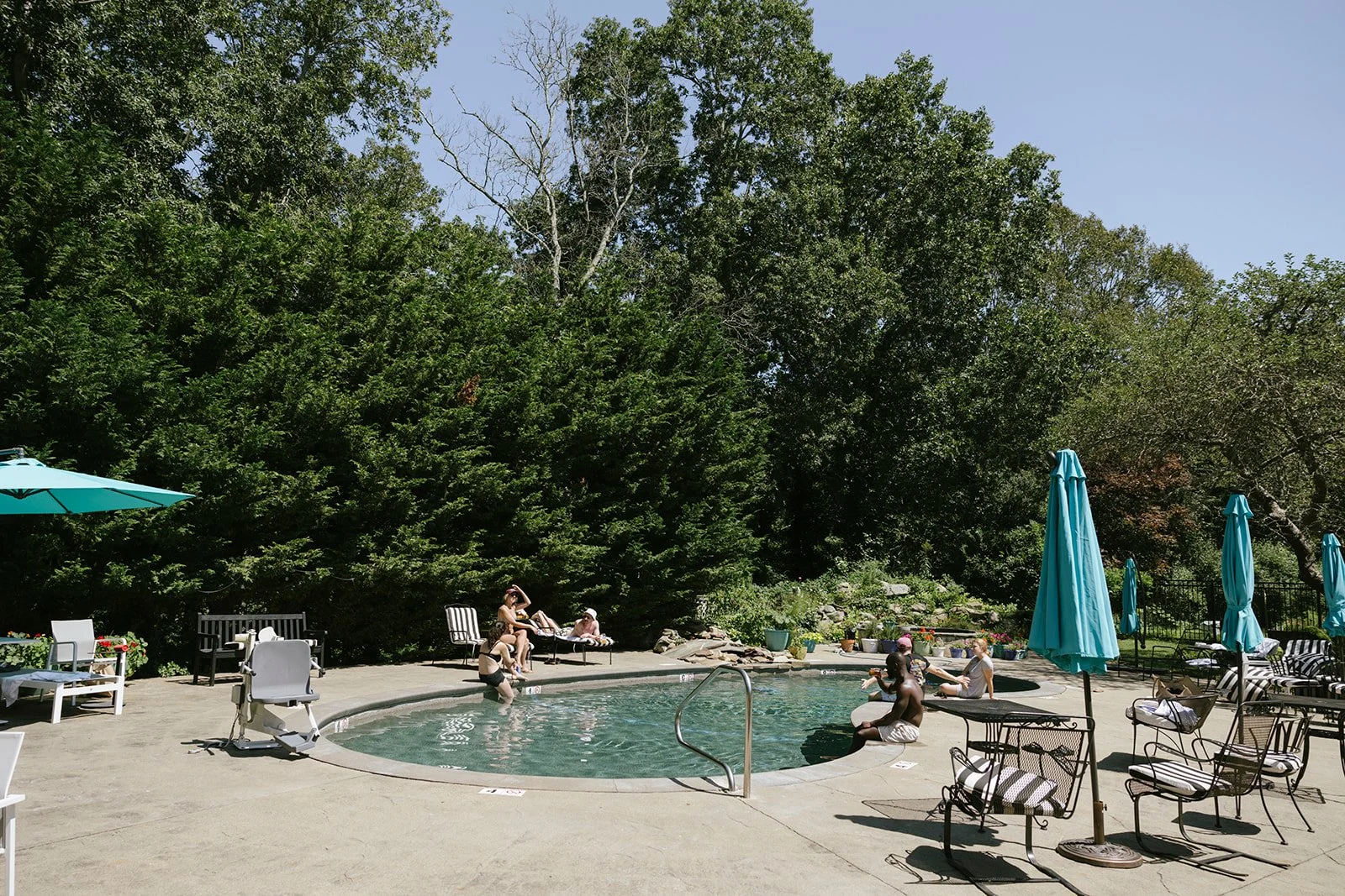 People relaxing by a the outdoor swimming pool at Lambert's Cove Inn on Martha's Vineyard surrounded by greenery and lawn chairs with umbrellas.