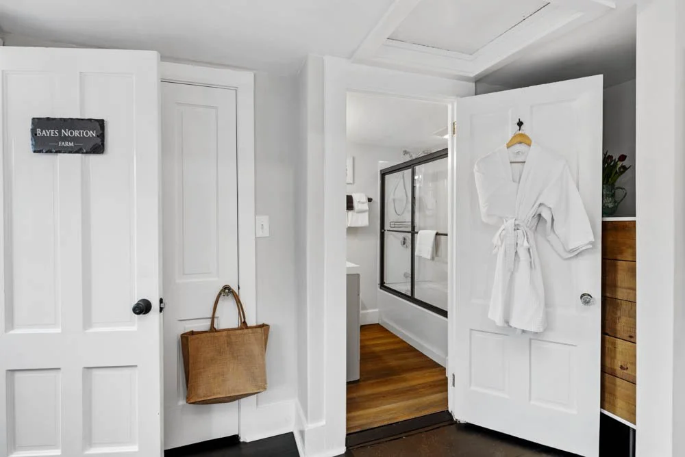 A bathroom at Lambert's Cove Inn with a partially open door hanging a white bathrobe. To the right, there is a wooden cabinet and a shower area with glass doors. The bathroom has wooden flooring. The inn has 15 guest rooms, a restaurant, and a bar. 