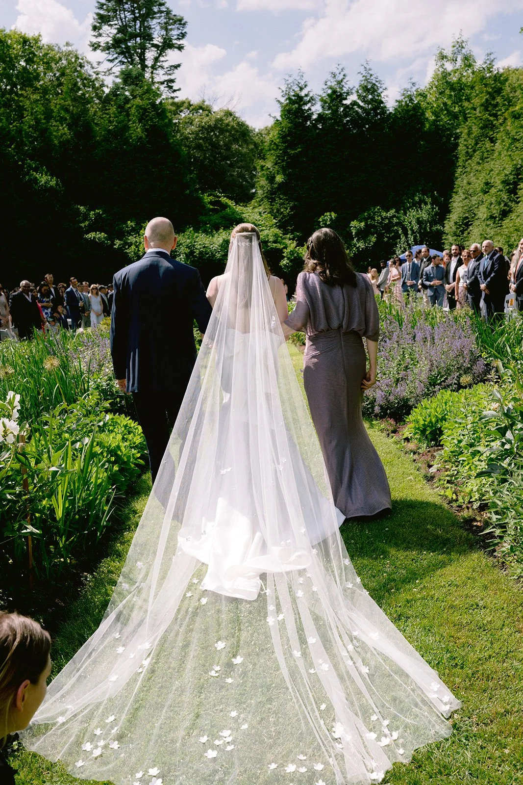 Bride walking down the aisle in a garden wedding at Lambert's Cove Inn on Martha's Vineyard, accompanied by her parents, as guests stand on either side on a sunny day.