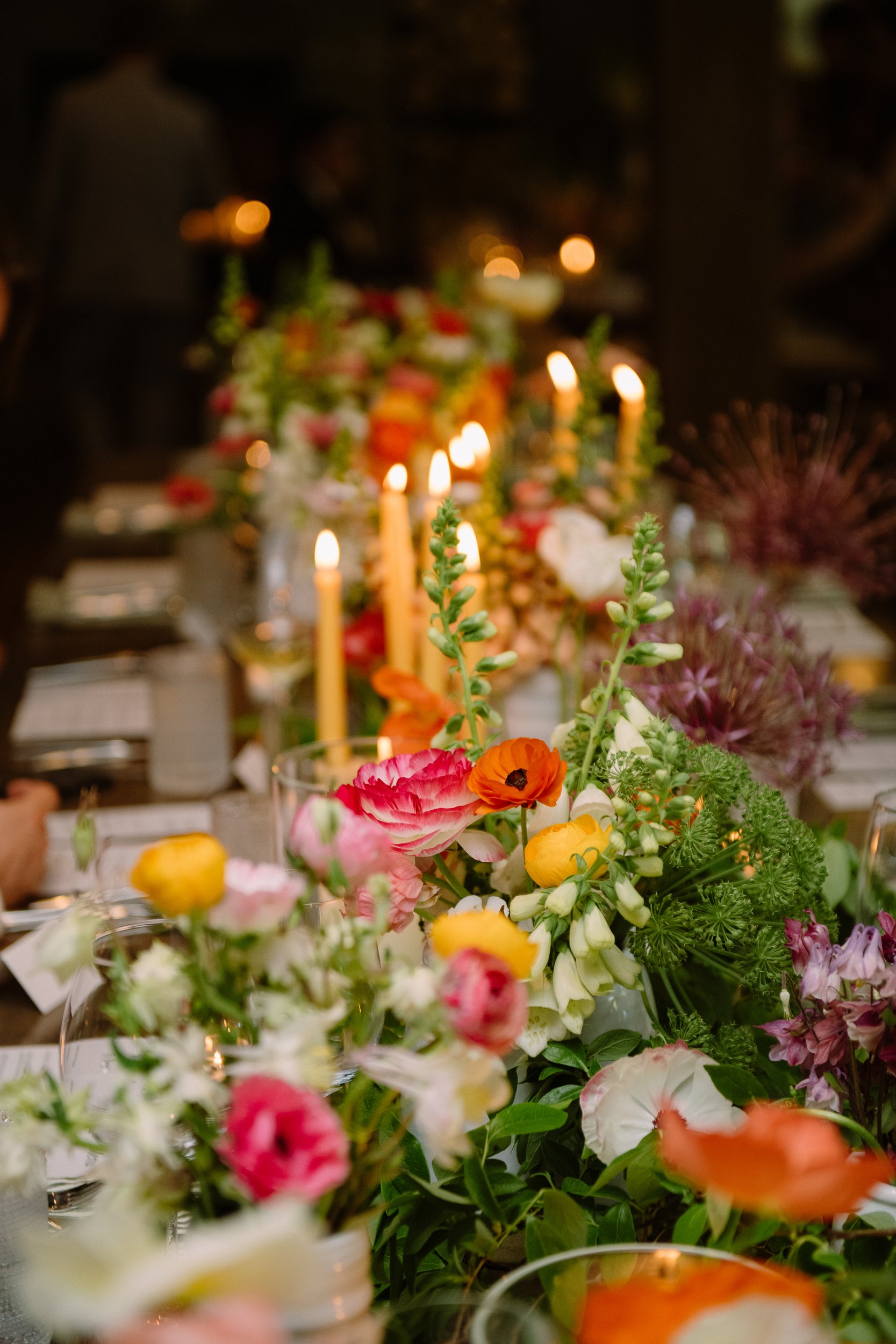 A floral table centerpiece with various colorful flowers including pink, white, yellow, and orange, set on a table with blurred background of candles and people in a dimly lit setting.