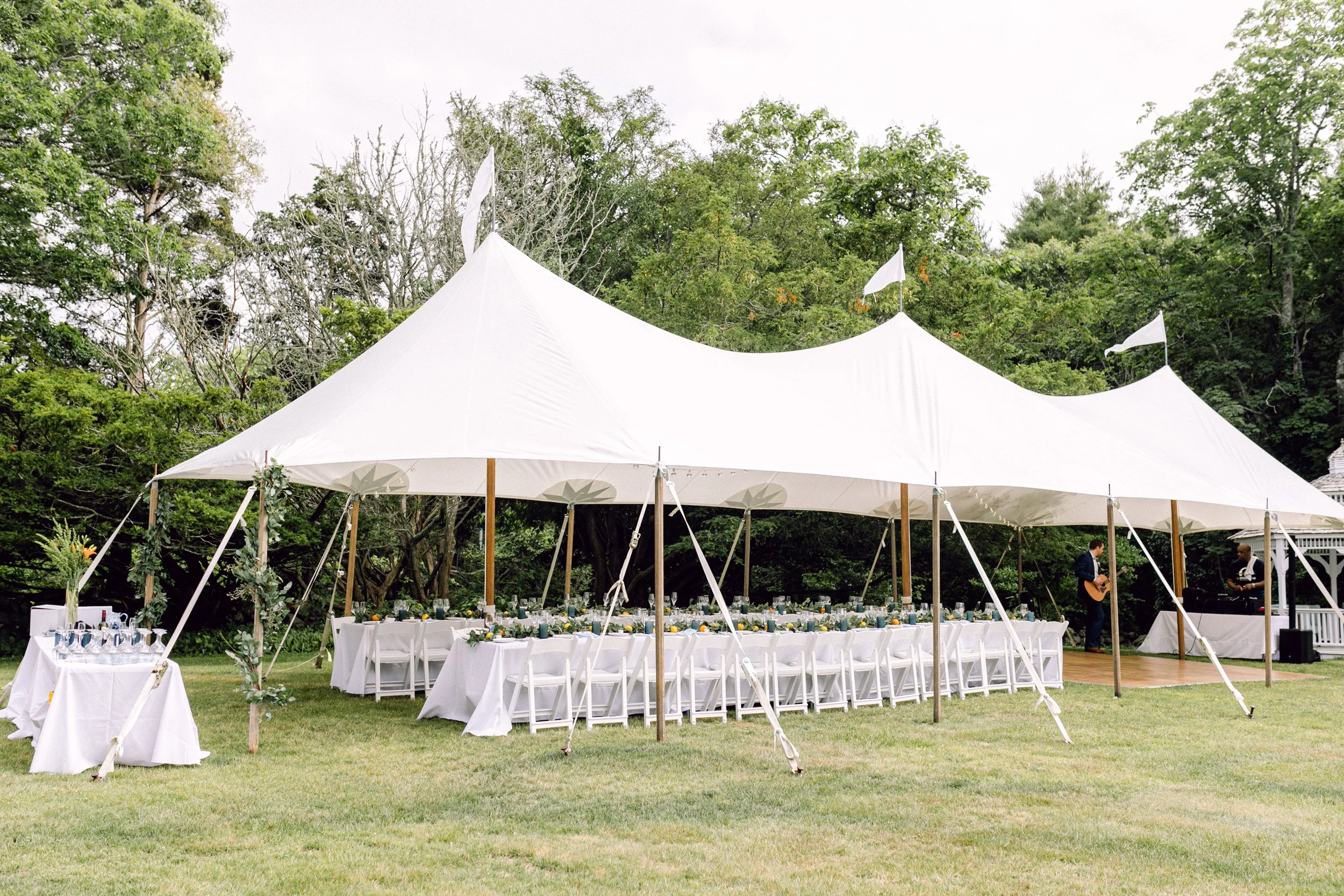 Large white wedding tent set up outdoors in a grassy area at Lambert's Cove Inn on Martha's Vineyard, surrounded by green trees, with tables and chairs underneath and two musicians playing guitar nearby.