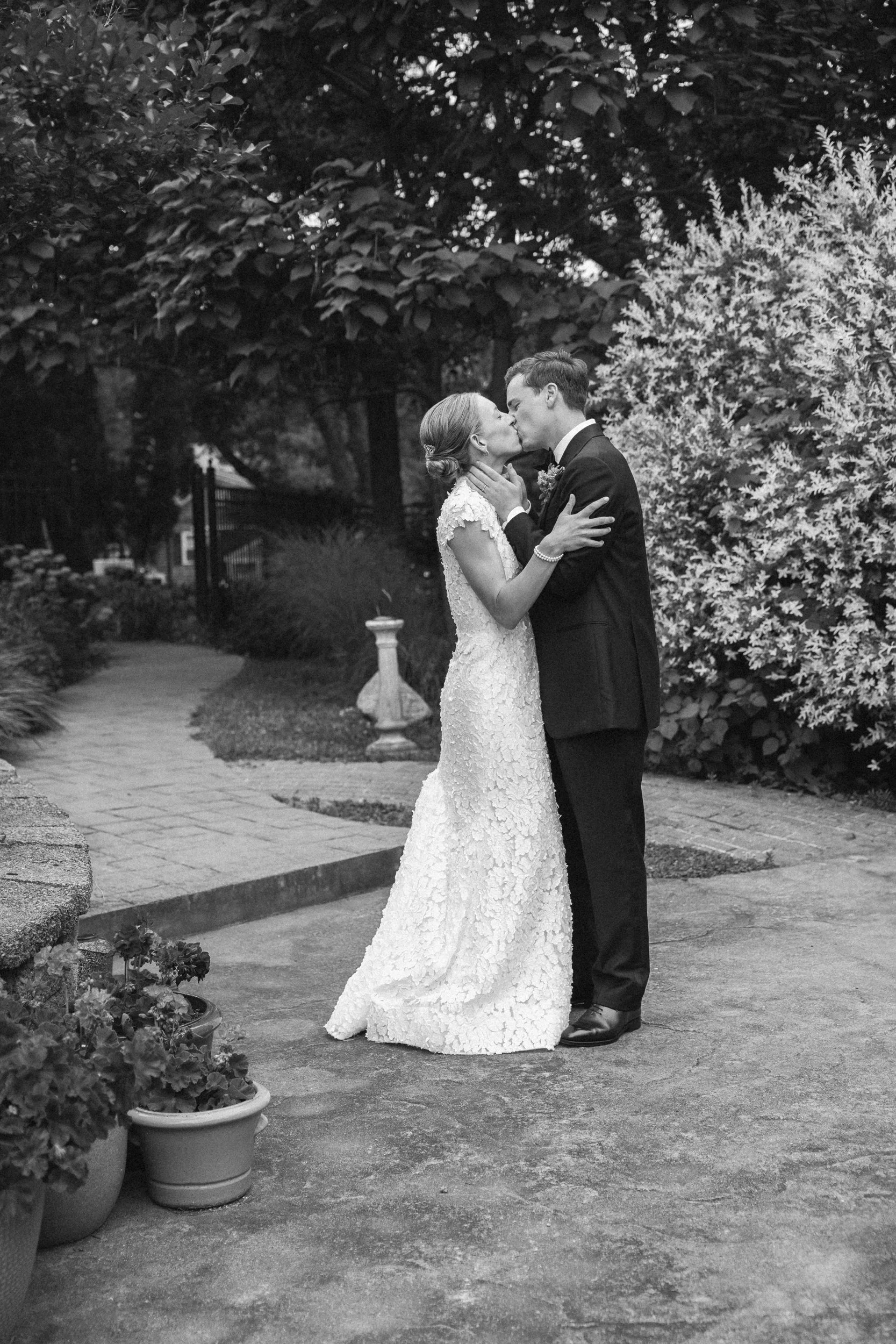 Black and white photograph of a bride and groom kissing outdoors. The bride wears a lace wedding gown and the groom is in a dark suit. They are standing on a paved path surrounded by bushes and trees during a wedding at Lambert's Cove Inn.